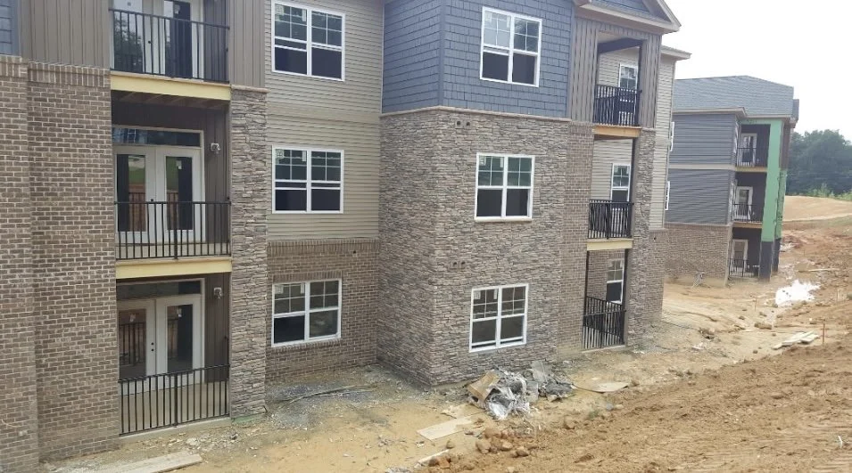 Under construction multi-story apartment building with brick and siding exterior, balconies with black railings, and unfinished ground area.