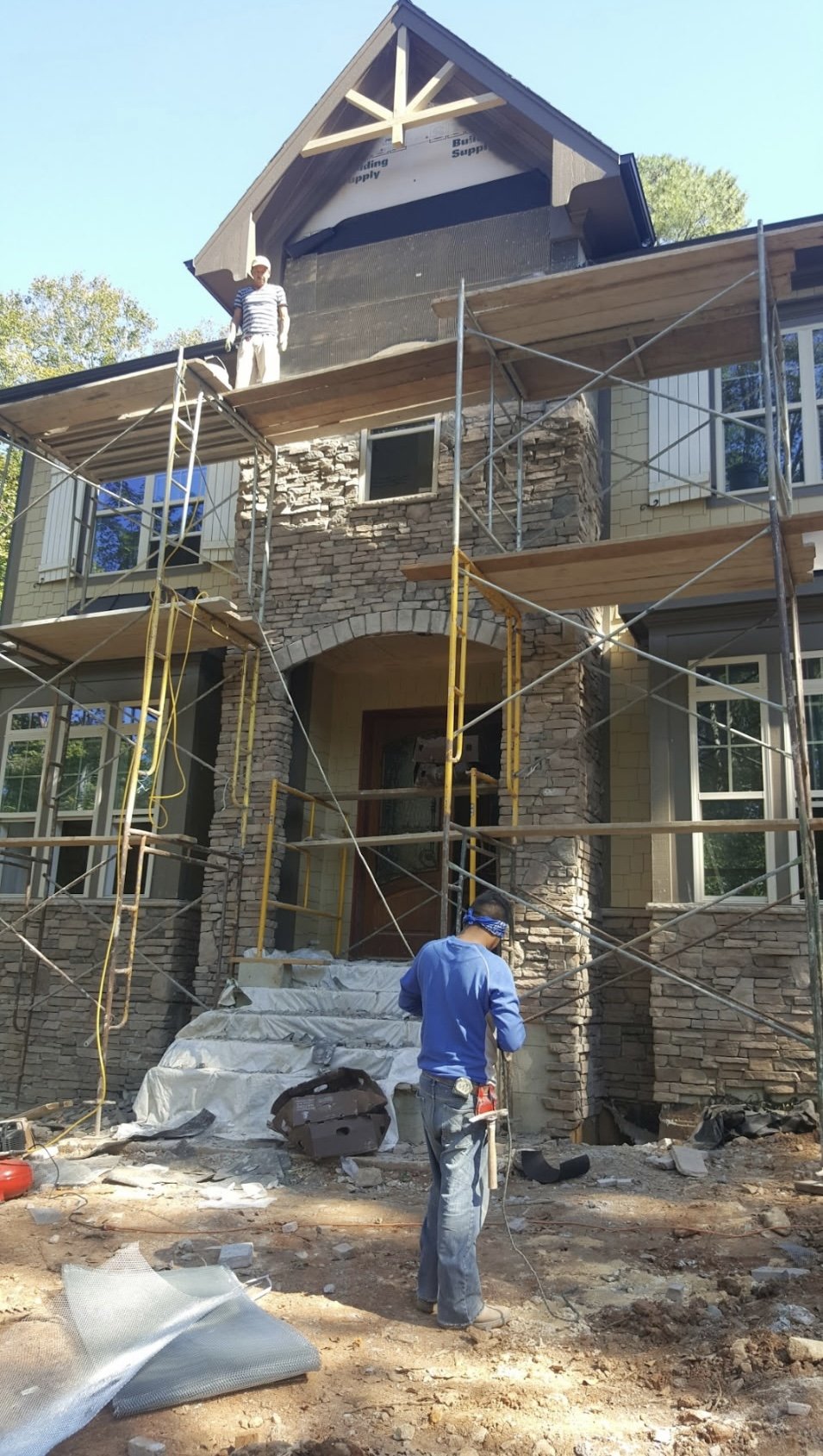 Construction workers building a multi-story house with stone and siding exterior, scaffolding around the front entrance, and a person working on the ground.