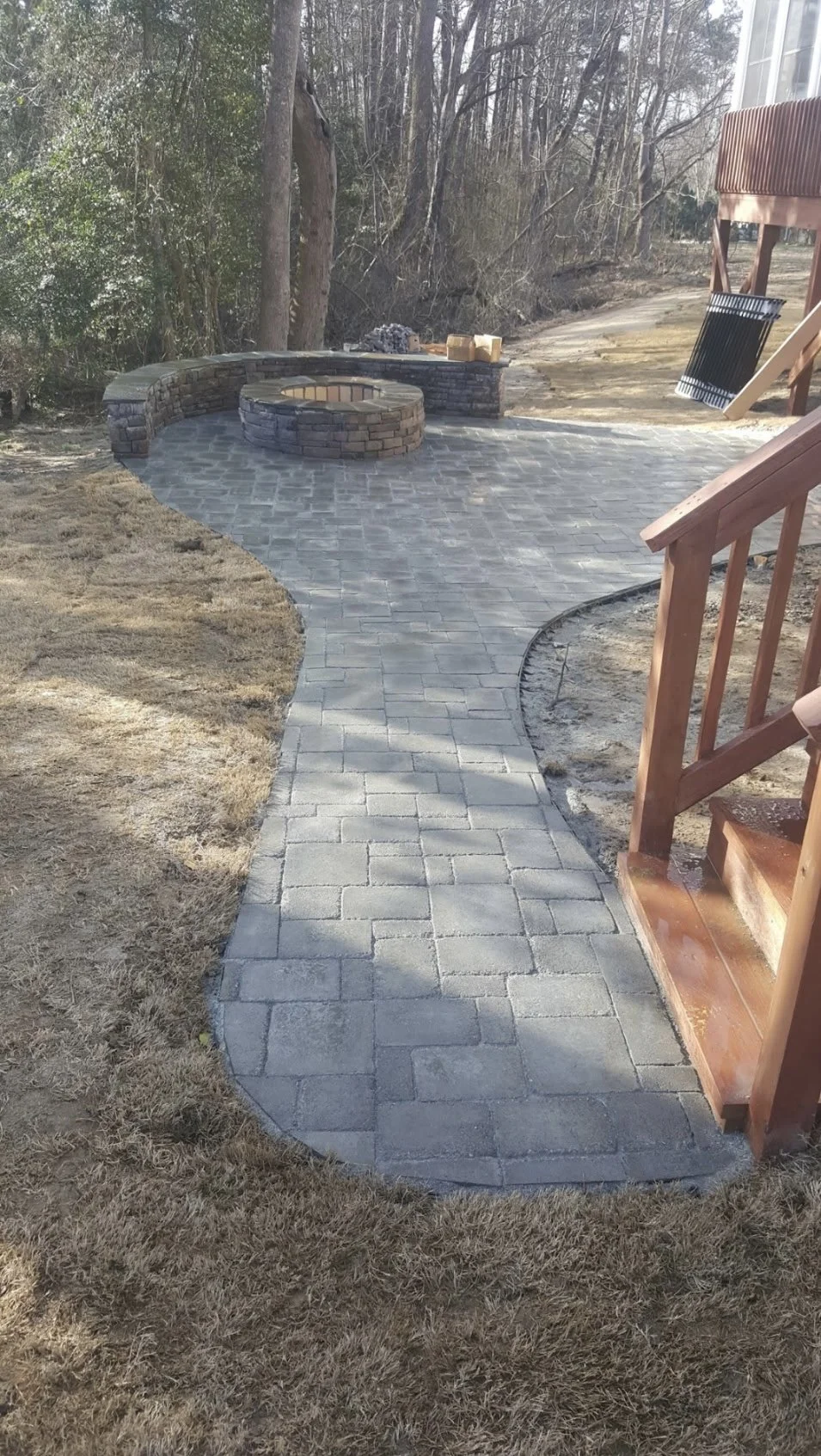 Backyard with a newly paved curved stone walkway leading to a small brick fire pit area with a brick wall and seating, surrounded by grass and trees.