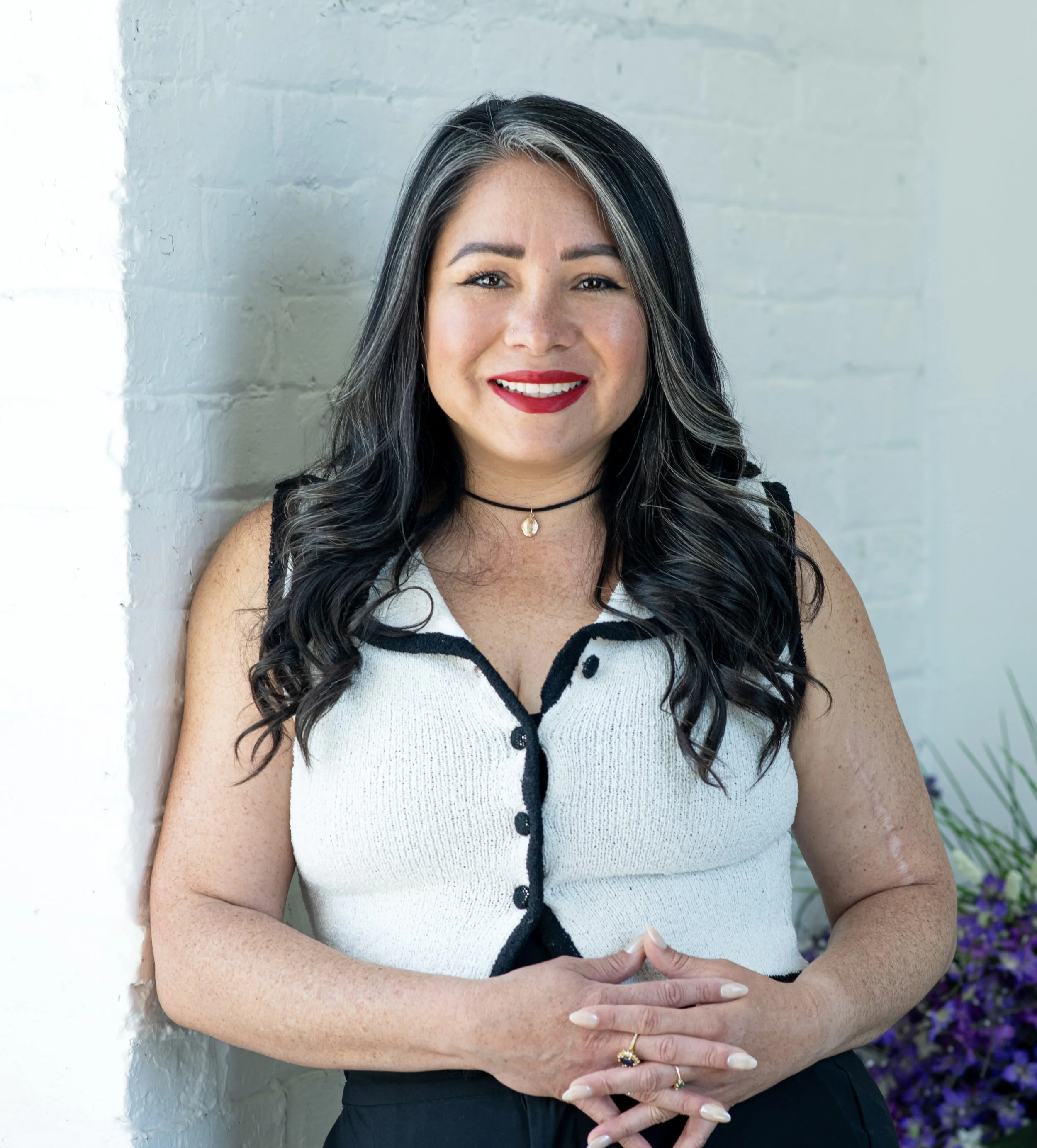 A woman with long black hair, wearing a sleeveless white top with black trim, smiling, standing against a white brick wall, with purple flowers visible on the right.