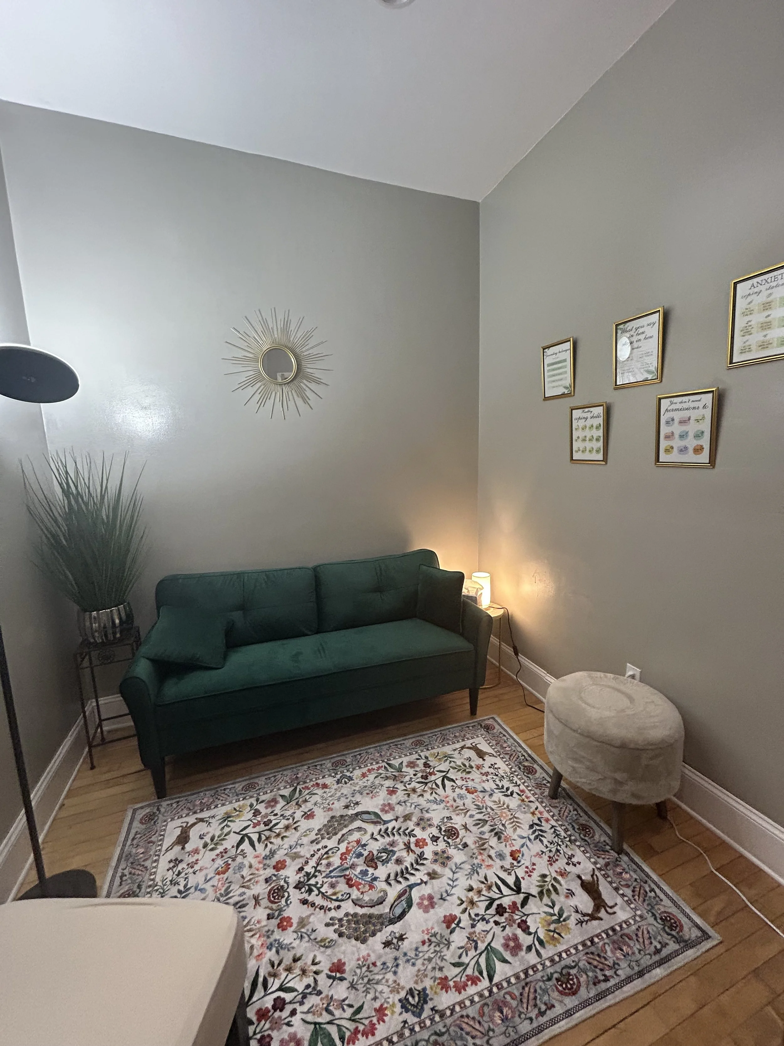 A cozy living room with a green sofa, a small beige ottoman, a patterned rug, a floor lamp, a table lamp, a large plant, and framed pictures on the wall.