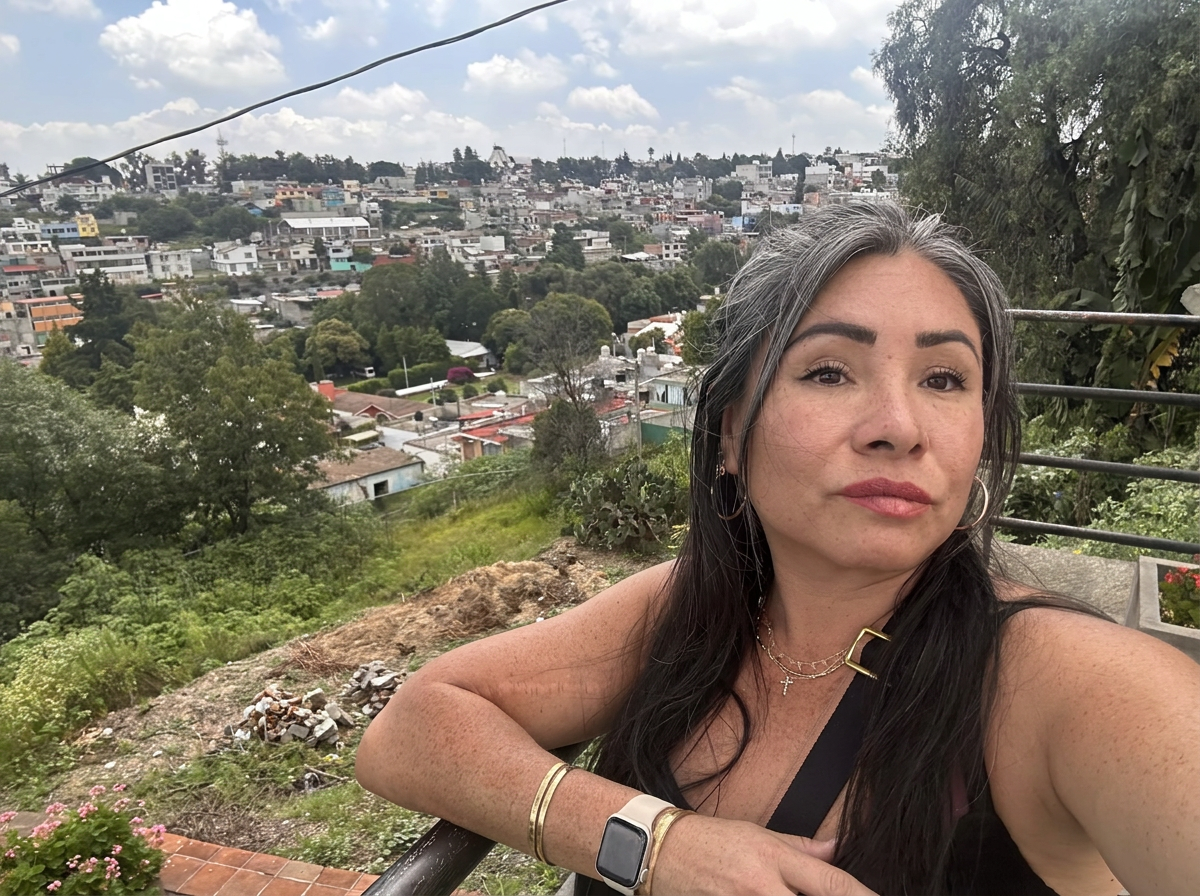 A woman with long dark hair and jewelry taking a selfie on a hillside with a view of a cityscape behind her, featuring buildings, trees, and a partly cloudy sky.