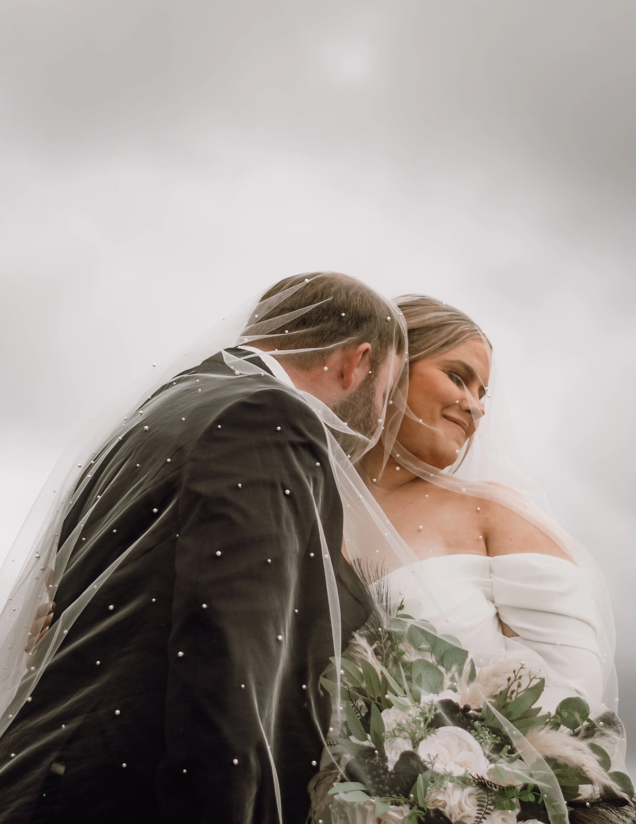 A bride and groom sharing a moment under a veil outdoors on a cloudy day, with the bride holding a bouquet of flowers.