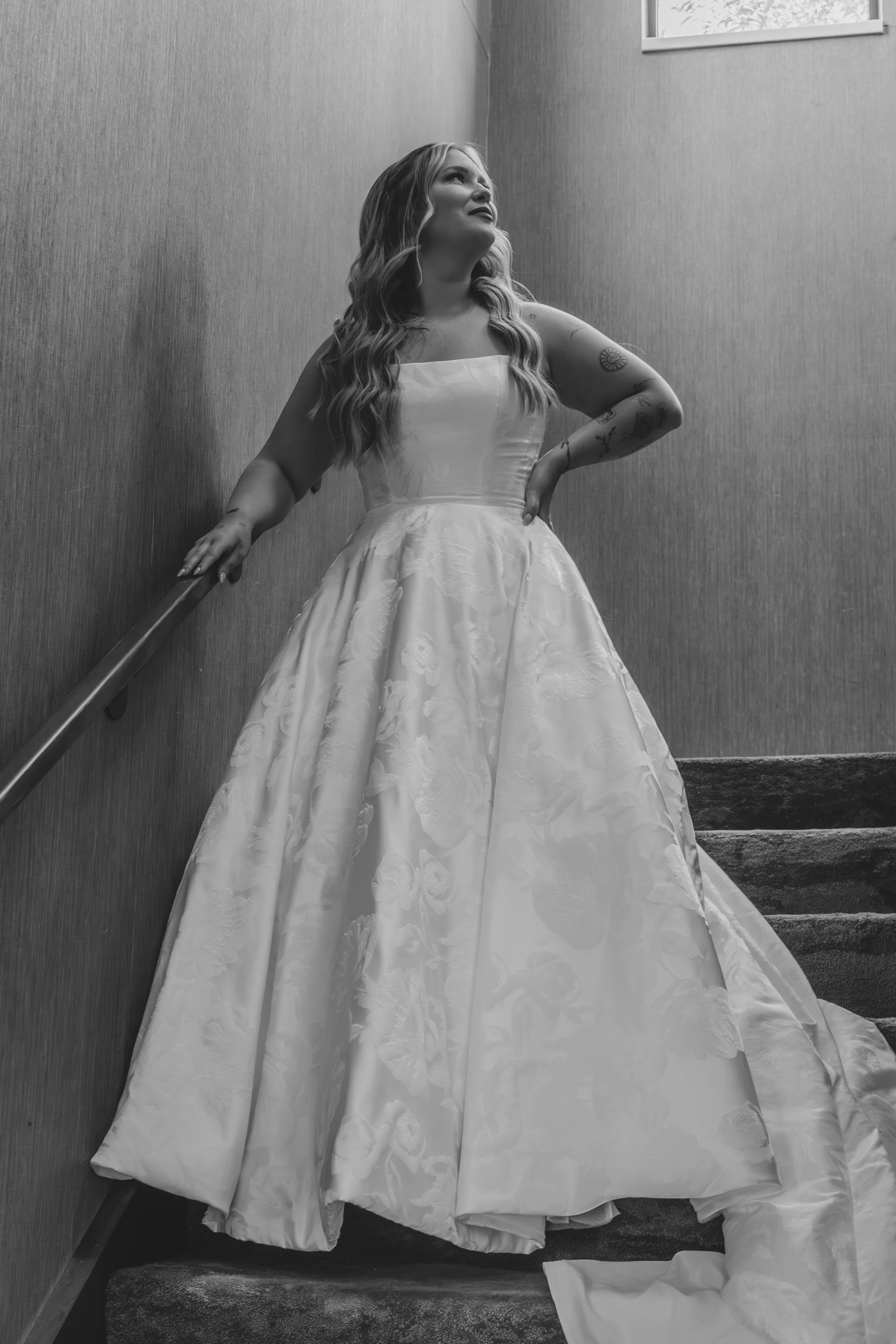 A woman in a wedding dress standing on stairs, looking up, with tattoos on her arm.
