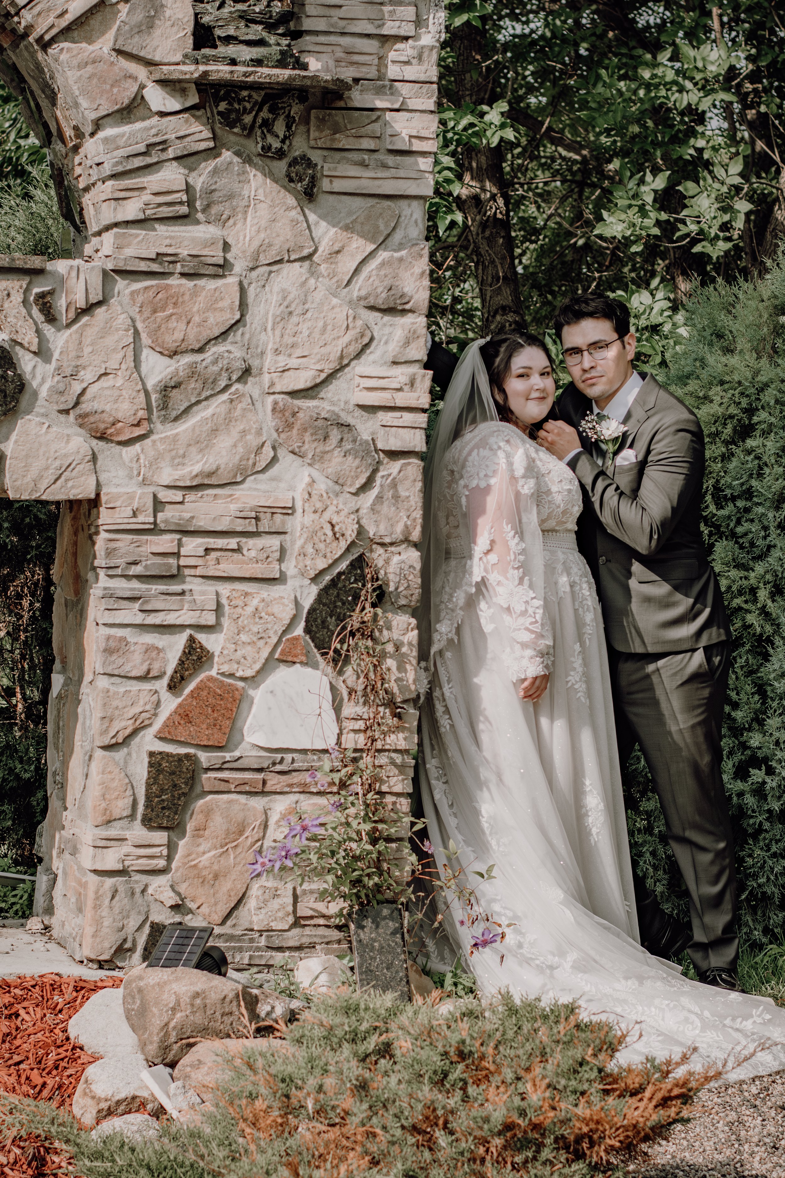 A bride and groom are posing outdoors, standing next to a stone wall with ivy and greenery in the background, during their wedding photo.