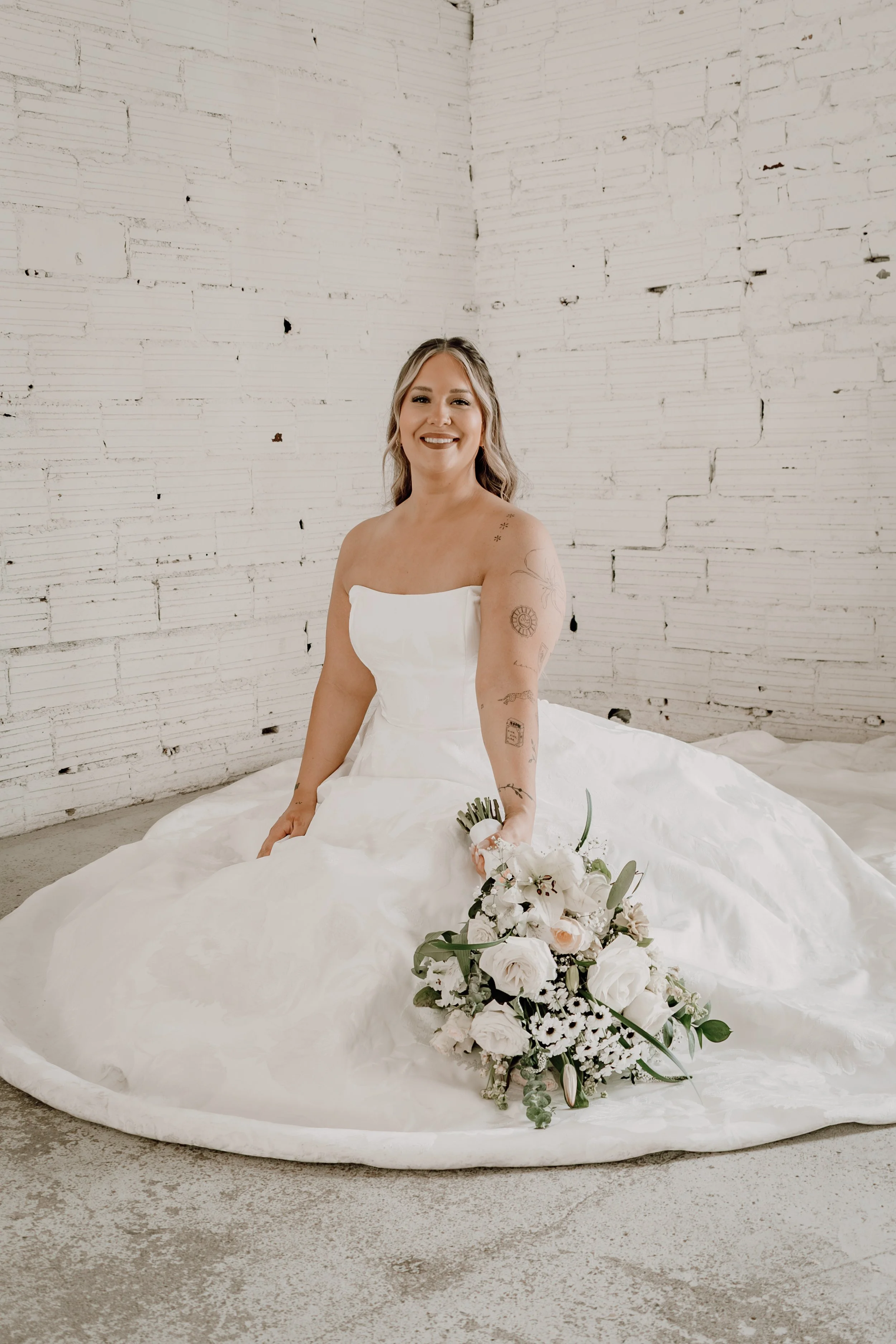 A smiling bride in a strapless white wedding dress, sitting on the floor against a white brick wall, holding a bouquet of white flowers.
