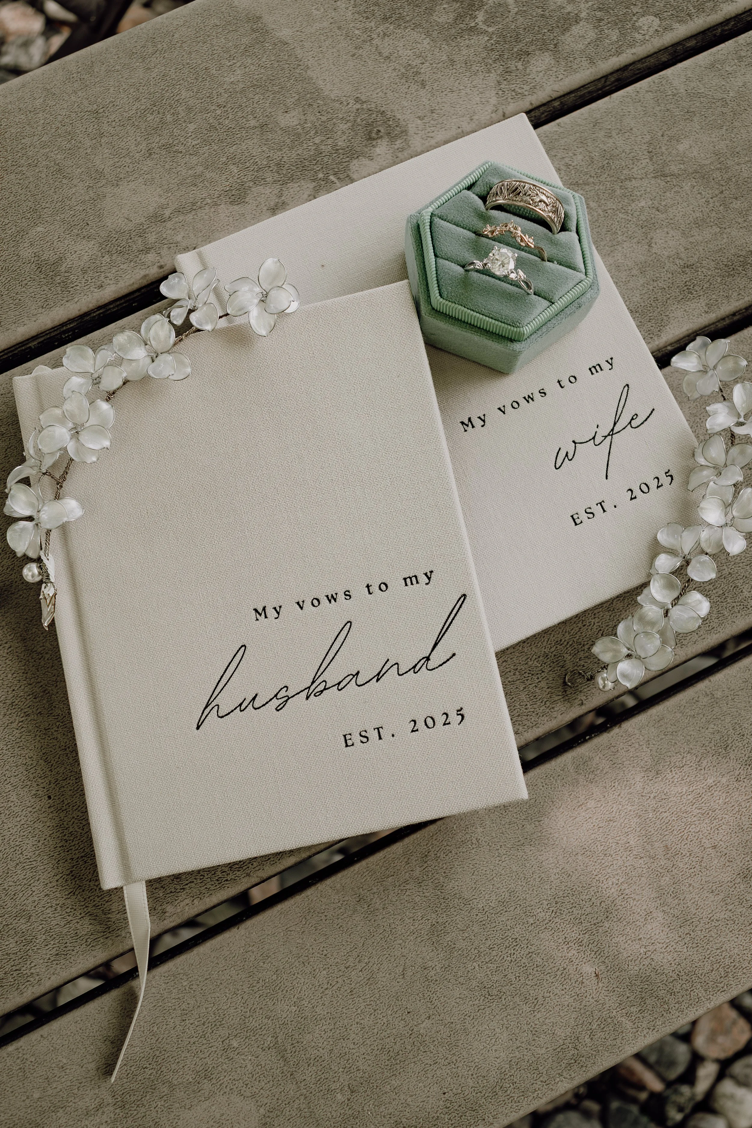Wedding vow booklets labeled "My vows to my husband" and "My vows to my wife," with the establishment year 2025, surrounded by a floral headpiece and a jewelry box with rings, on a bench.