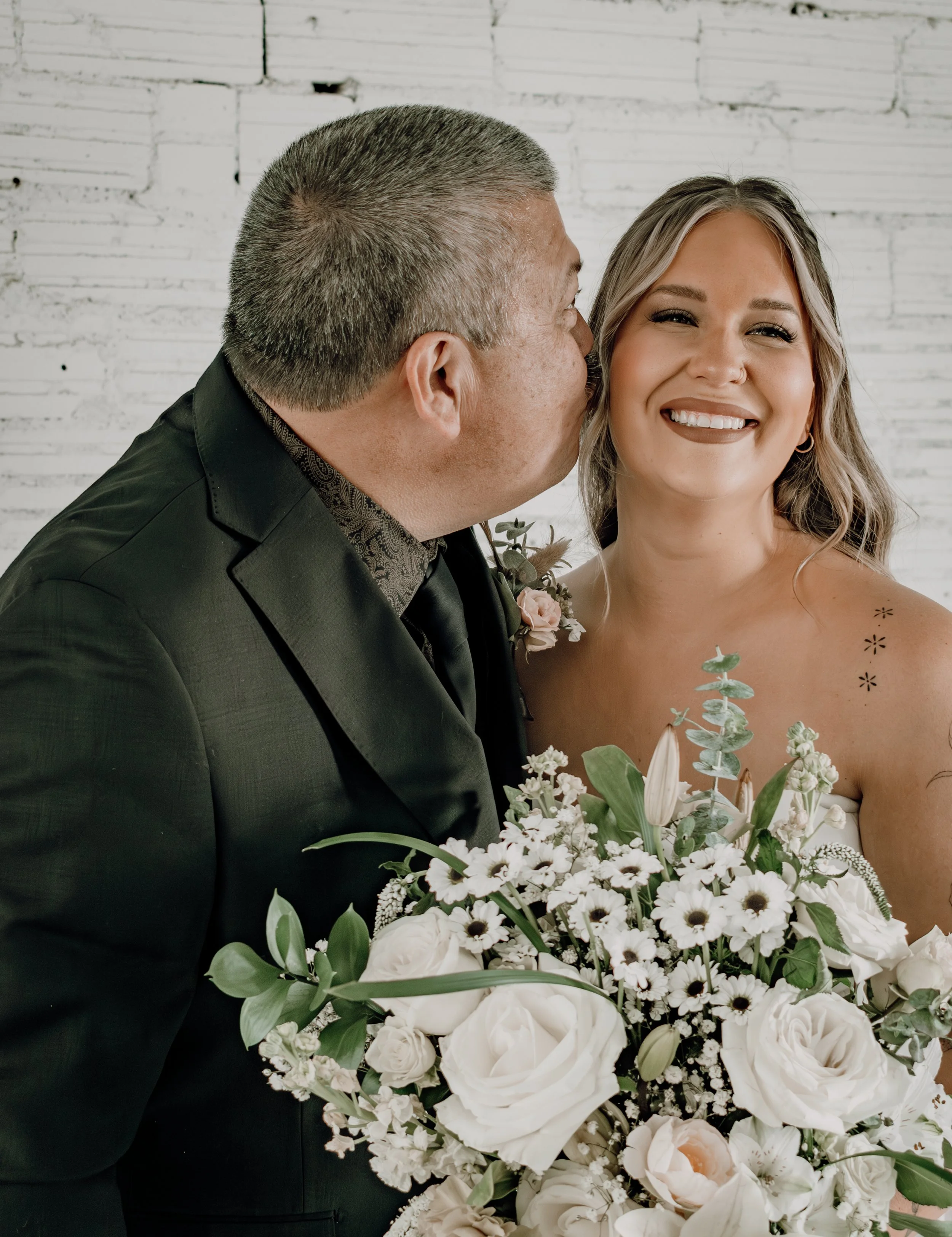 A groom kissing a bride on the cheek during their wedding, holding a large bouquet of white flowers, in front of a white brick wall.