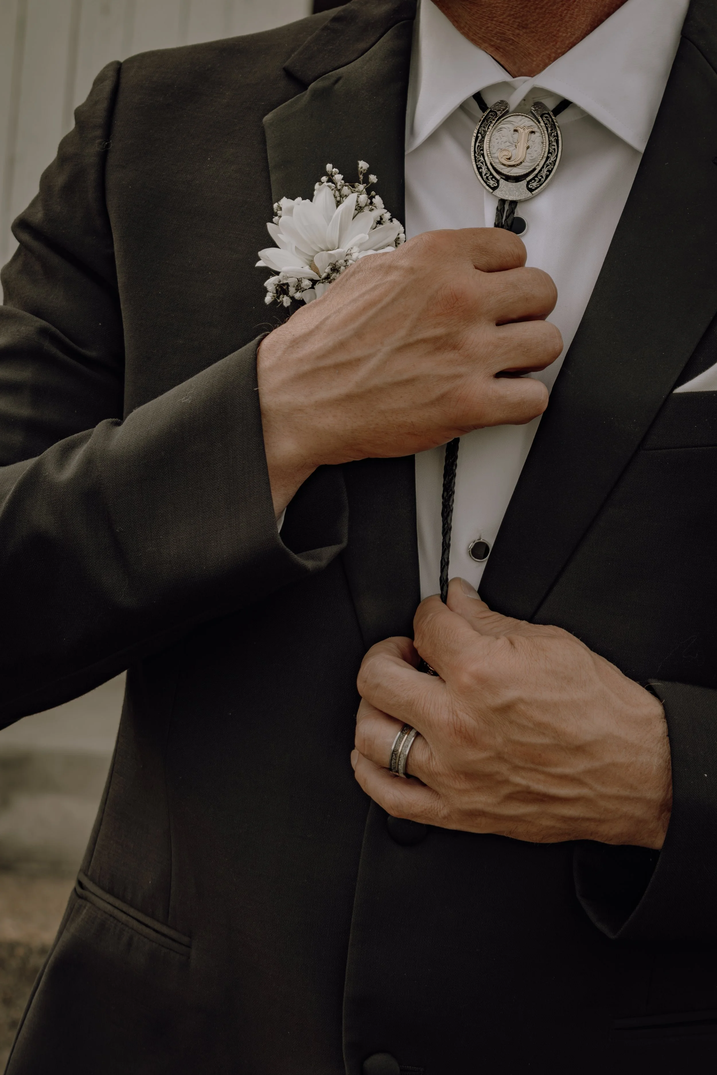 Close-up of a man adjusting his black suit jacket. He has a boutonnière with white flowers on his left lapel and is wearing a bolo tie with a silver clasp featuring a 'J'. He is also wearing a wedding band on his left ring finger.