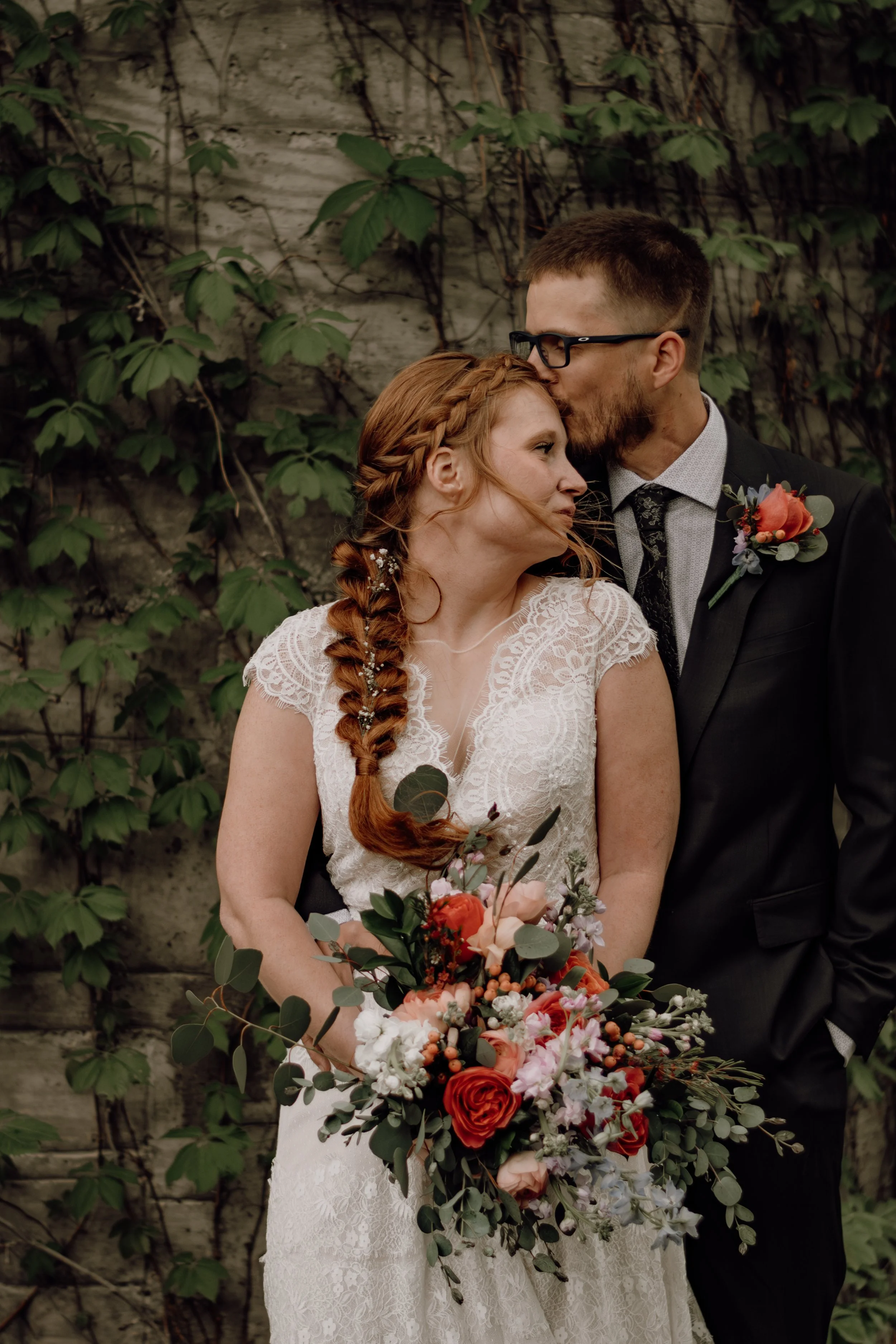 A bride and groom share a romantic moment, with the groom kissing the bride’s forehead. The bride holds a large bouquet of colorful flowers, and the groom is wearing a suit with a boutonniere. They are standing outdoors against a backdrop of green iv