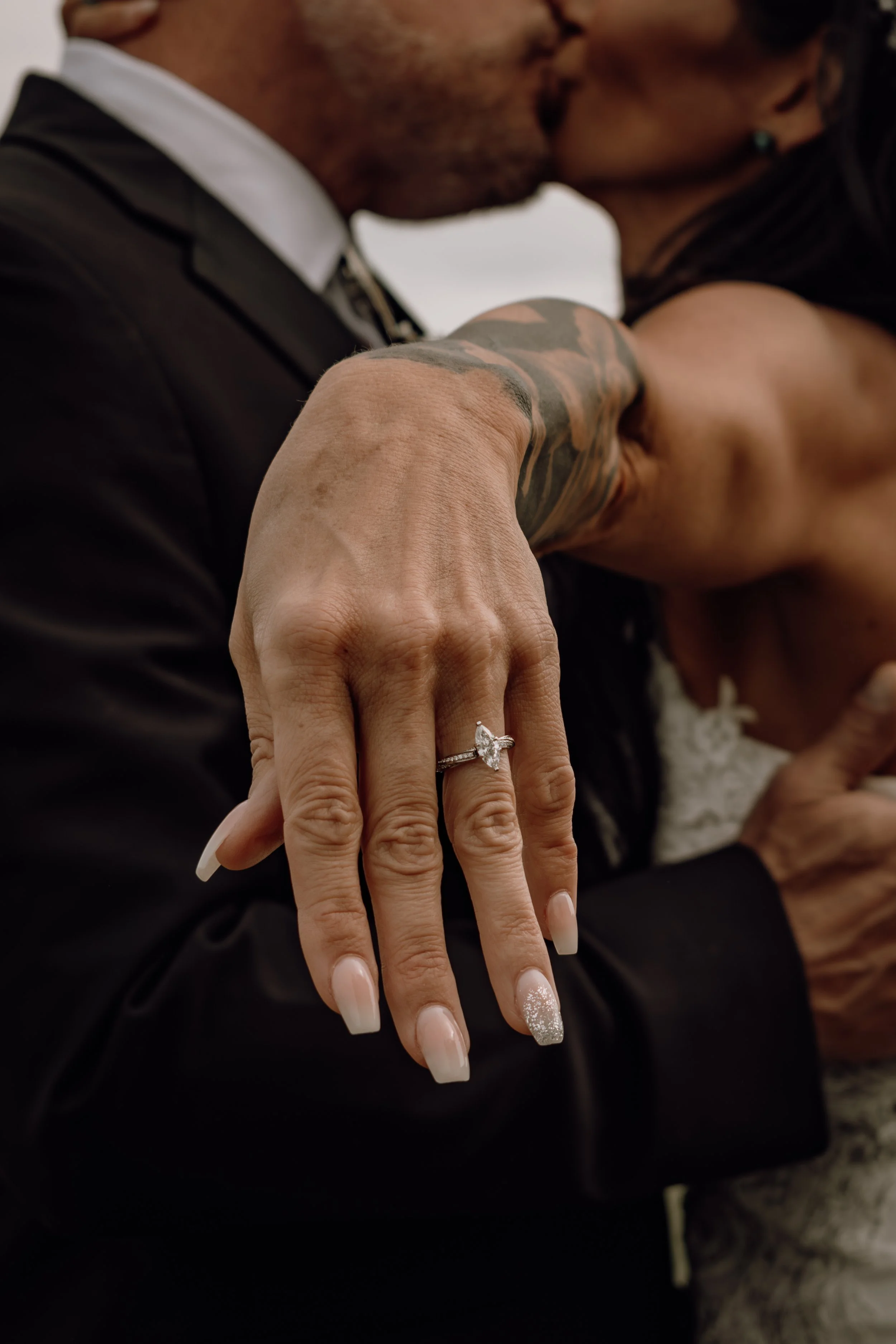 A close-up of a woman's hand showing a wedding ring with a large heart-shaped diamond. The woman has a manicure with French tips and glitter accents. She is being kissed on the lips by a man, who has tattooed arms and is dressed in a black suit and w