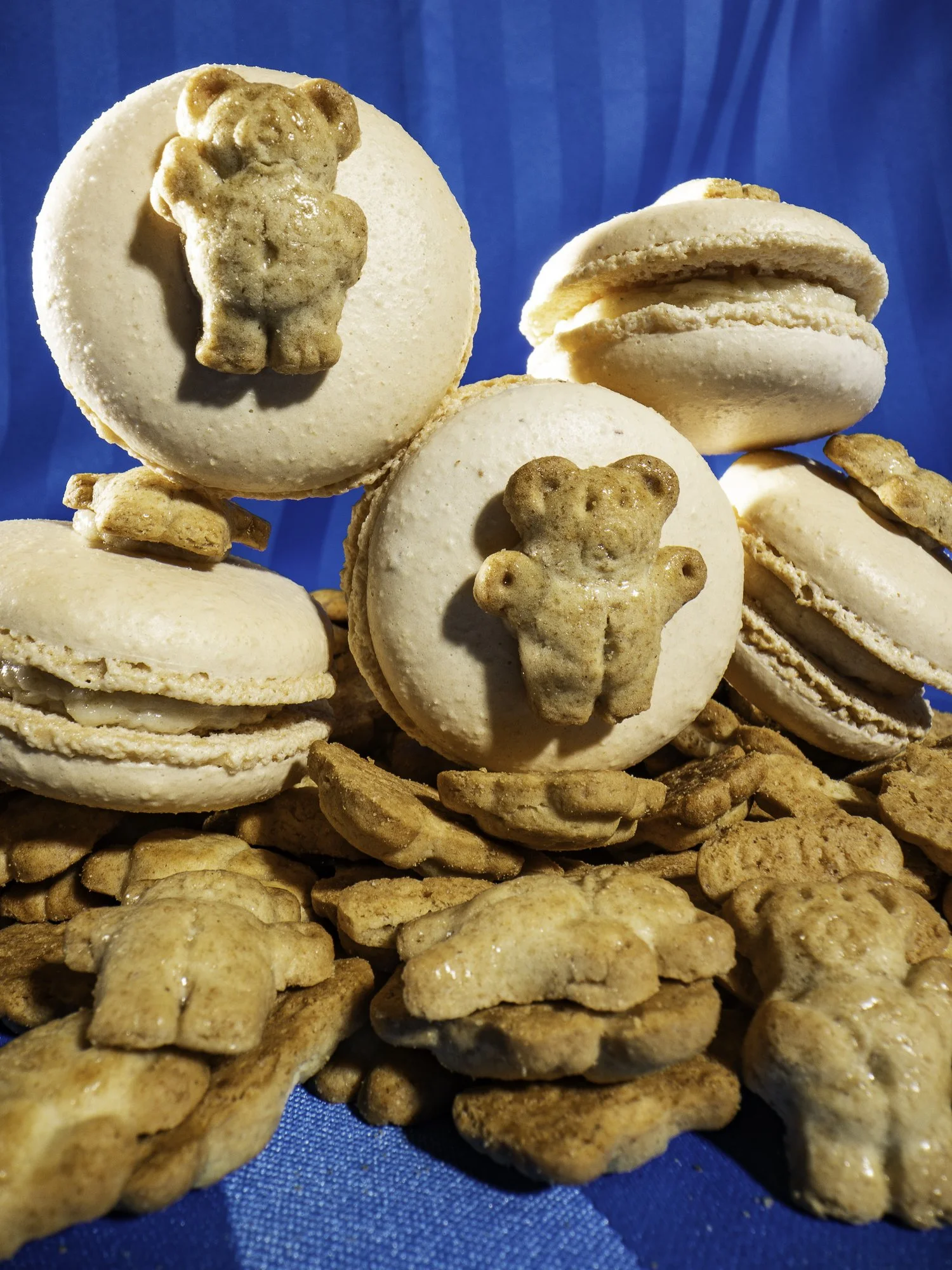 Cream-colored macarons with teddy bear-shaped cookies on top, surrounded by small bear-shaped cookies, all on a blue surface.