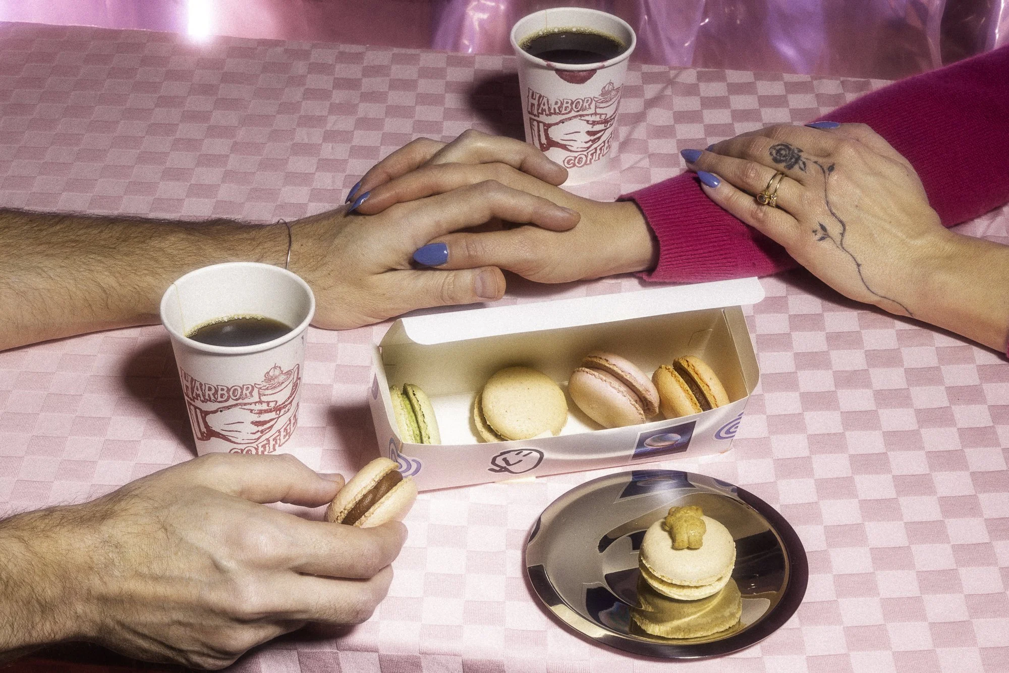 Two people with painted nails and tattoos, holding hands across a pink checkered table. On the table are two cups of coffee, a box of assorted macarons, and a small plate with a macaron topped with a teddy bear-shaped cookie.