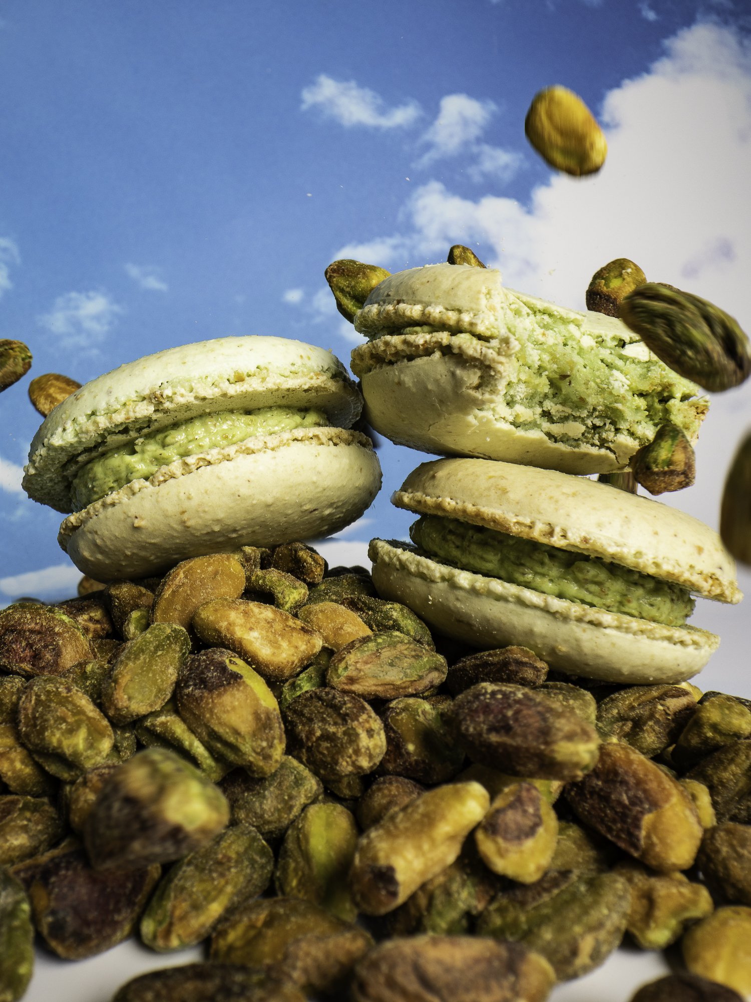Close-up of pistachio macarons with whole pistachios and broken shells, some falling, against a blue sky with clouds.