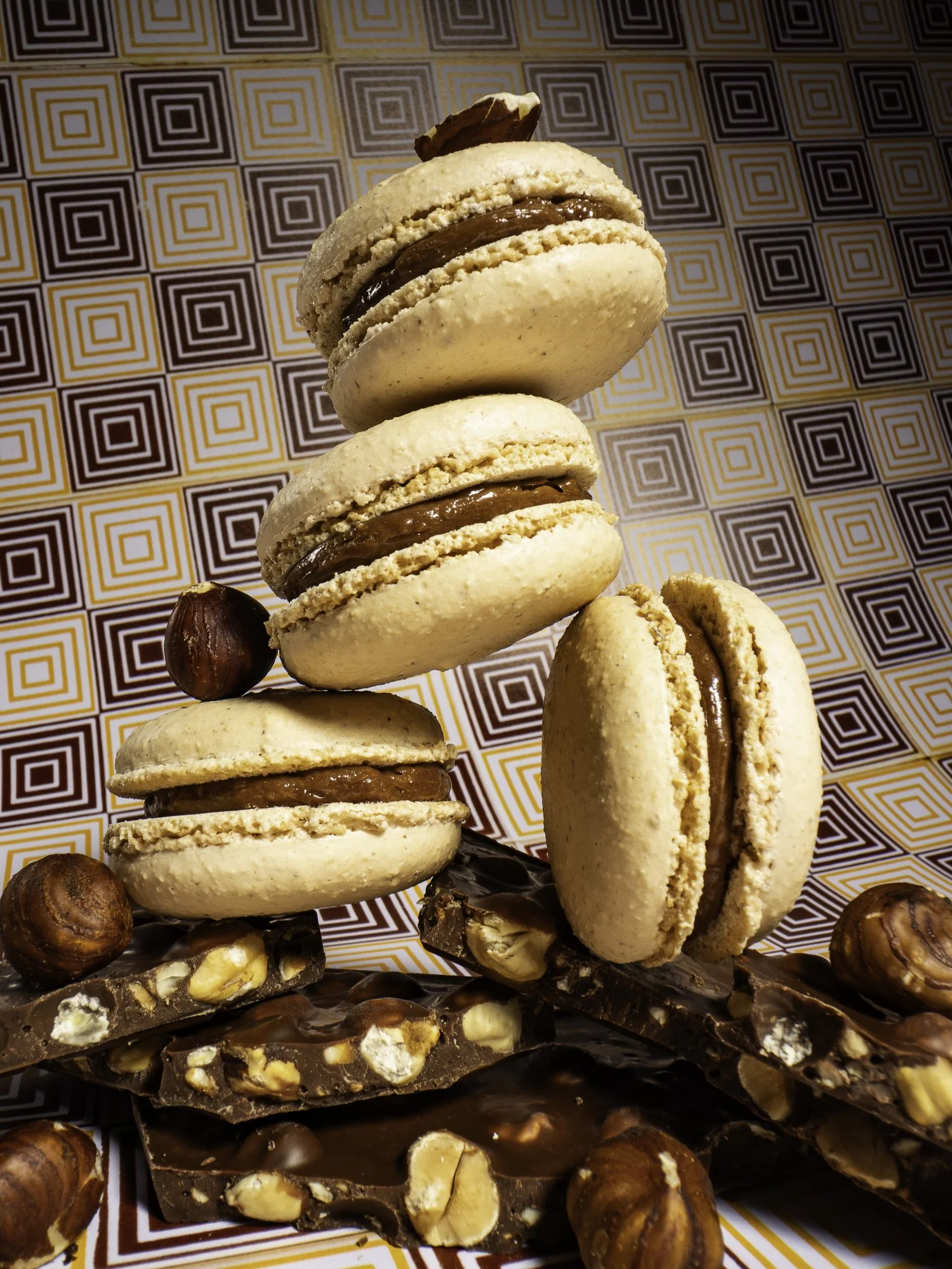 Stack of four French macarons with chocolate filling, surrounded by hazelnuts and chocolate almond bark, on a patterned background.
