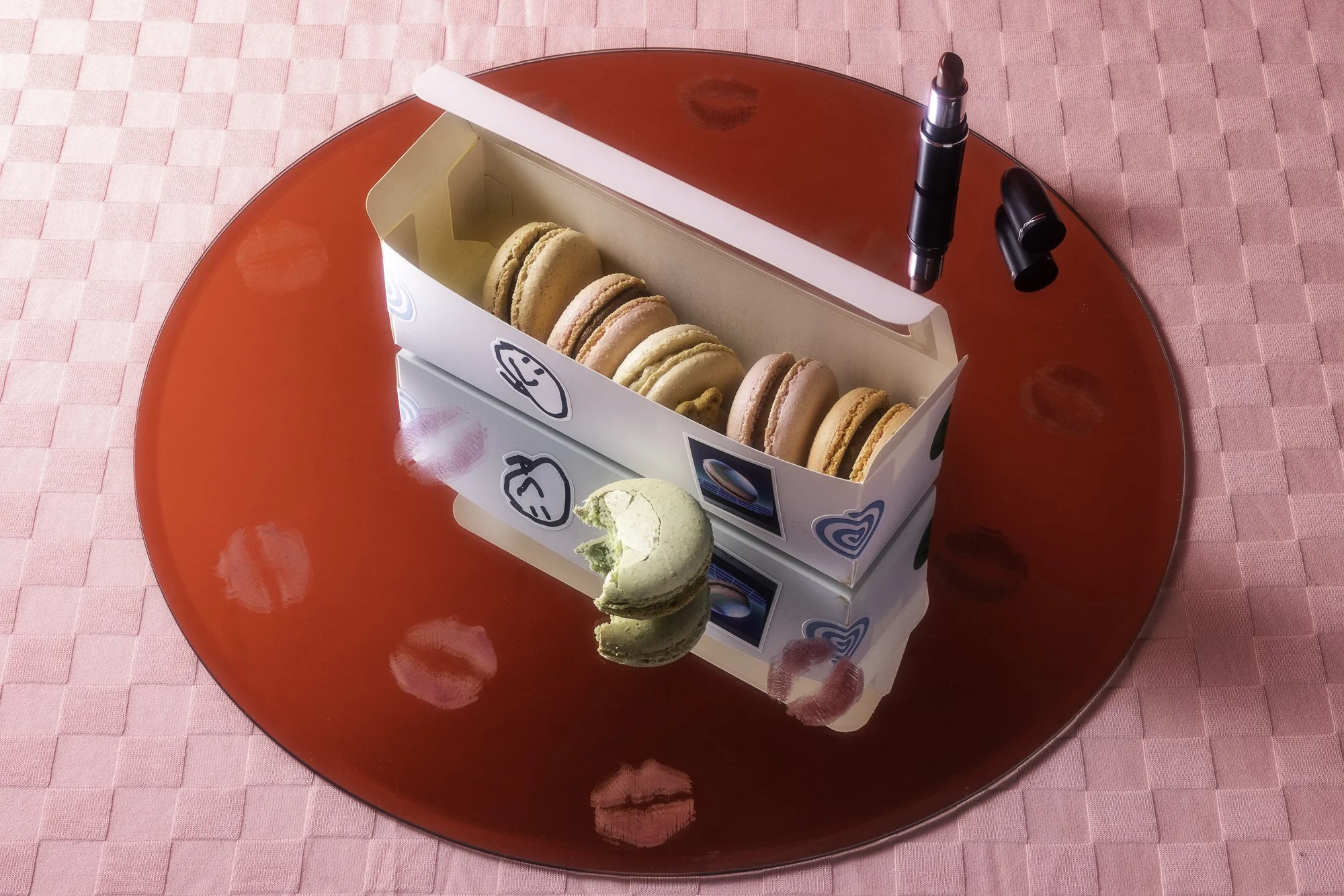 A box of macarons on a red round plate with lipstick and lip gloss nearby, on a pink tablecloth.