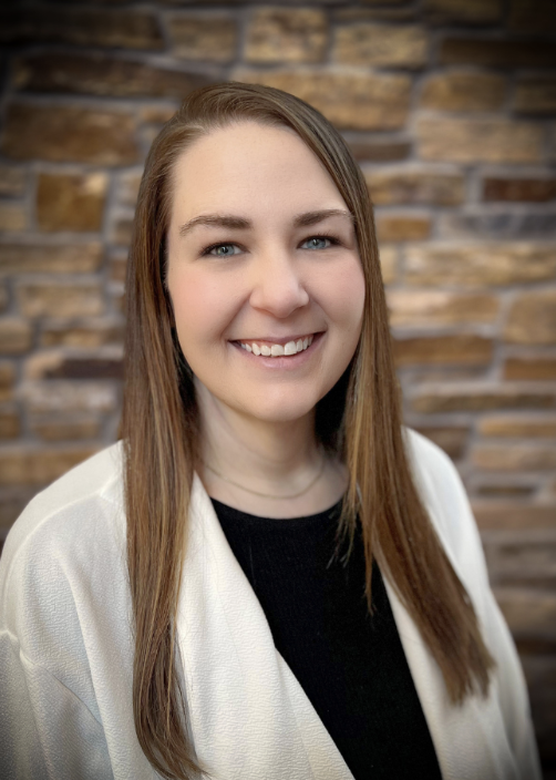 Kyla Conway, a white woman with long straight brown hair and blue eyes smiling at the camera with a brick wall in the backdrop. 