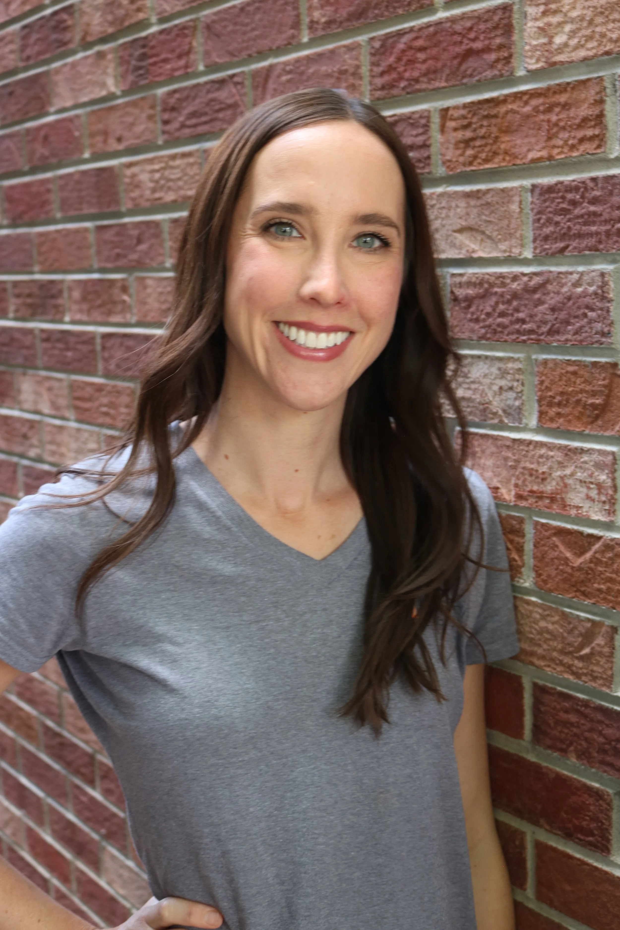 Dr Kimberlie Landers, a white woman with long wavy brown hear, blue eyes and smiling directly at the camera with a brick wall in the background. 