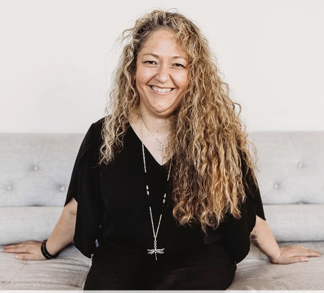 Liliana Baylon, a Latina woman with long curly brown hair smiling at the camera, wearing a loose black top and a dragonfly necklace. 