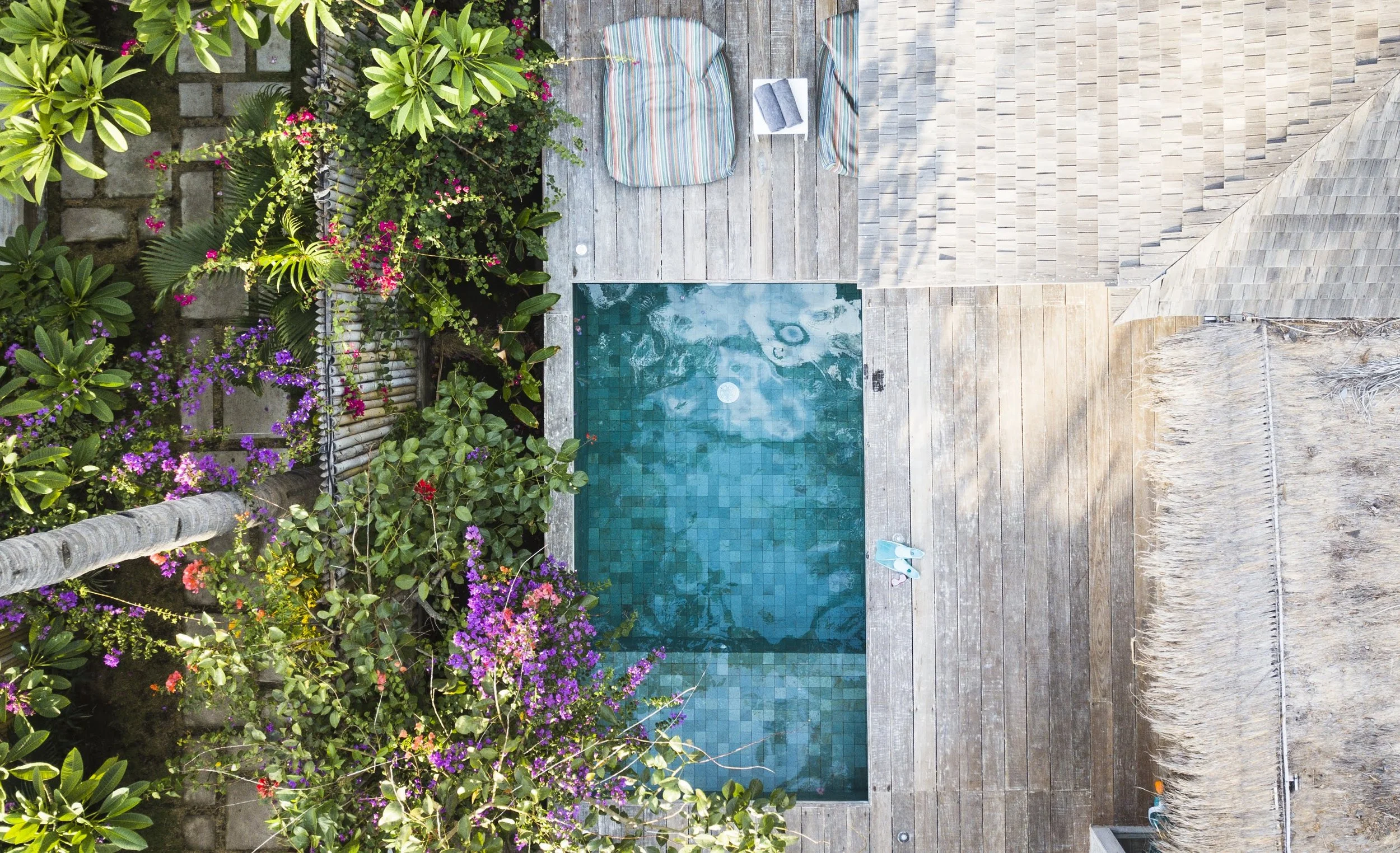Piscine rectangulaire avec un revêtement en carreaux bleus, entourée d'une terrasse en bois, plantes fleuries et végétation tropicale sur le côté gauche, et une structure en toit de chaume à droite. Ile des Gilis, Indonésie 
