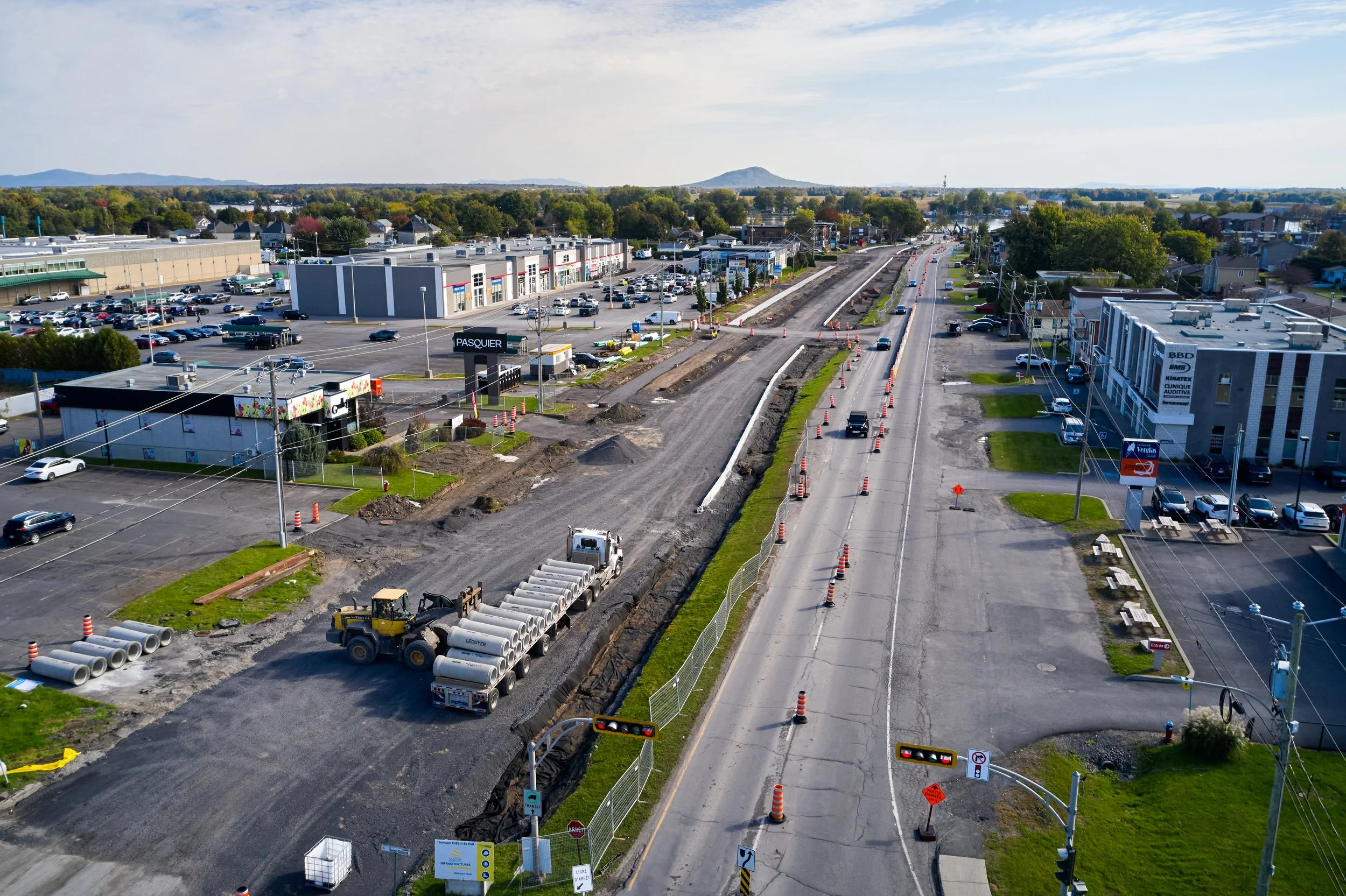 Travaux routiers en cours sur une avenue urbaine avec des cônes de signalisation, des machines de construction et des routes partiellement fermées. Magasins et parkings visibles en arrière-plan.