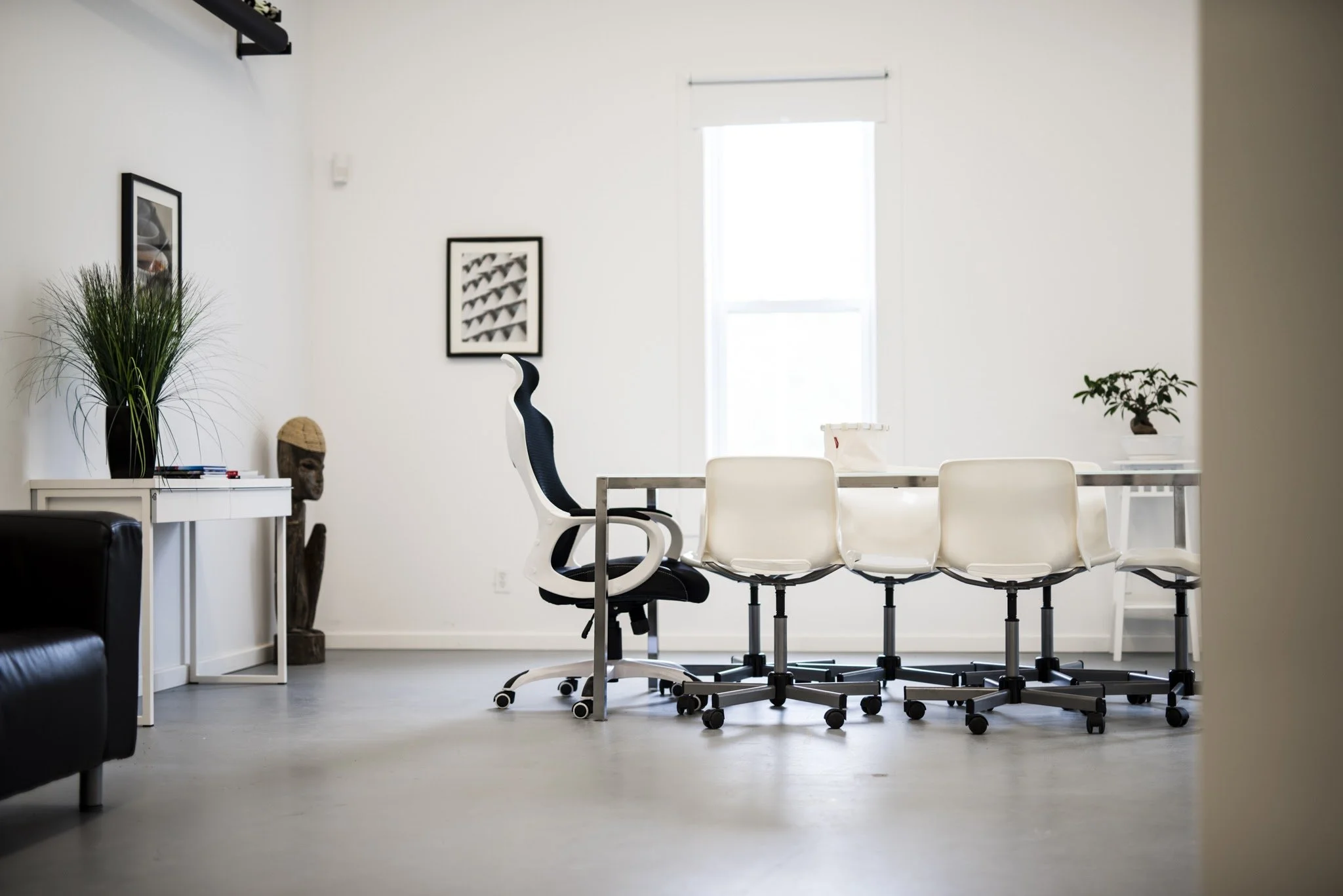 Salle de réunion ou espace de travail avec une grande table blanche, plusieurs chaises blanches sur roulettes disposées autour, une grande fenêtre laissant entrer la lumière, décor avec des plantes et des œuvres d'art
