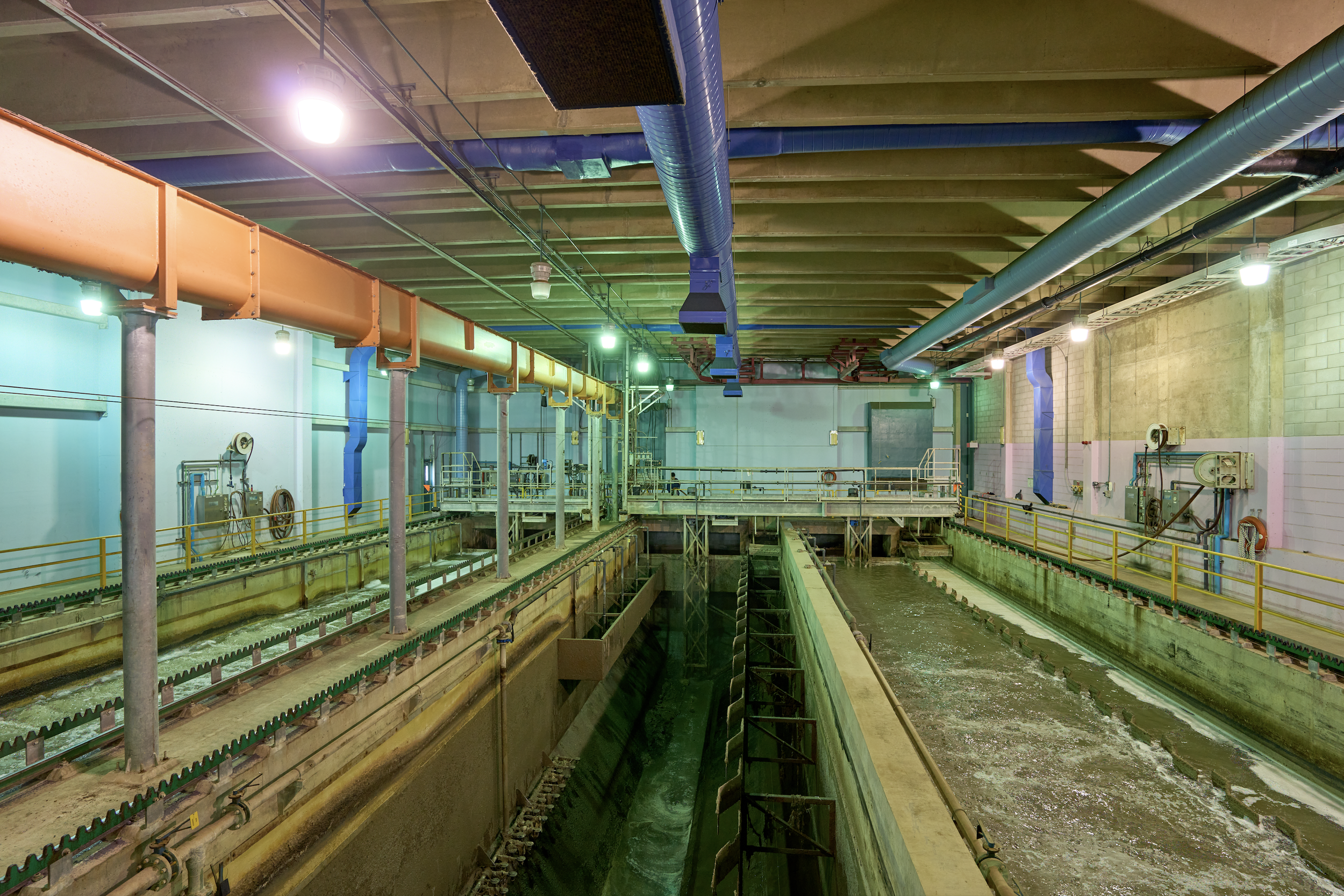 Salle industrielle avec des canalisations colorées, une fosse ou canalisation de traitement et des murs en béton, éclairée par des lumières suspendues.