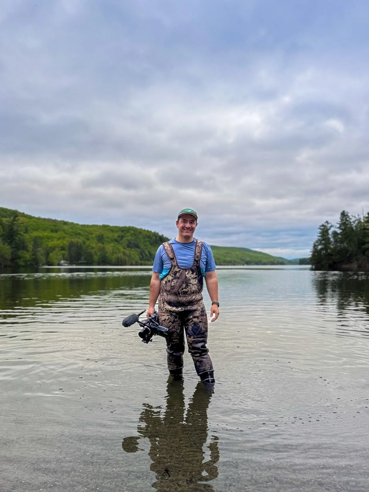 Un caméraman debout dans l'eau d'une rivière, tenant une caméra FX6 Sony portant des bottes de pêche et une tenue camouflage, souriant, avec un paysage de forêt et un ciel nuageux en arrière-plan.