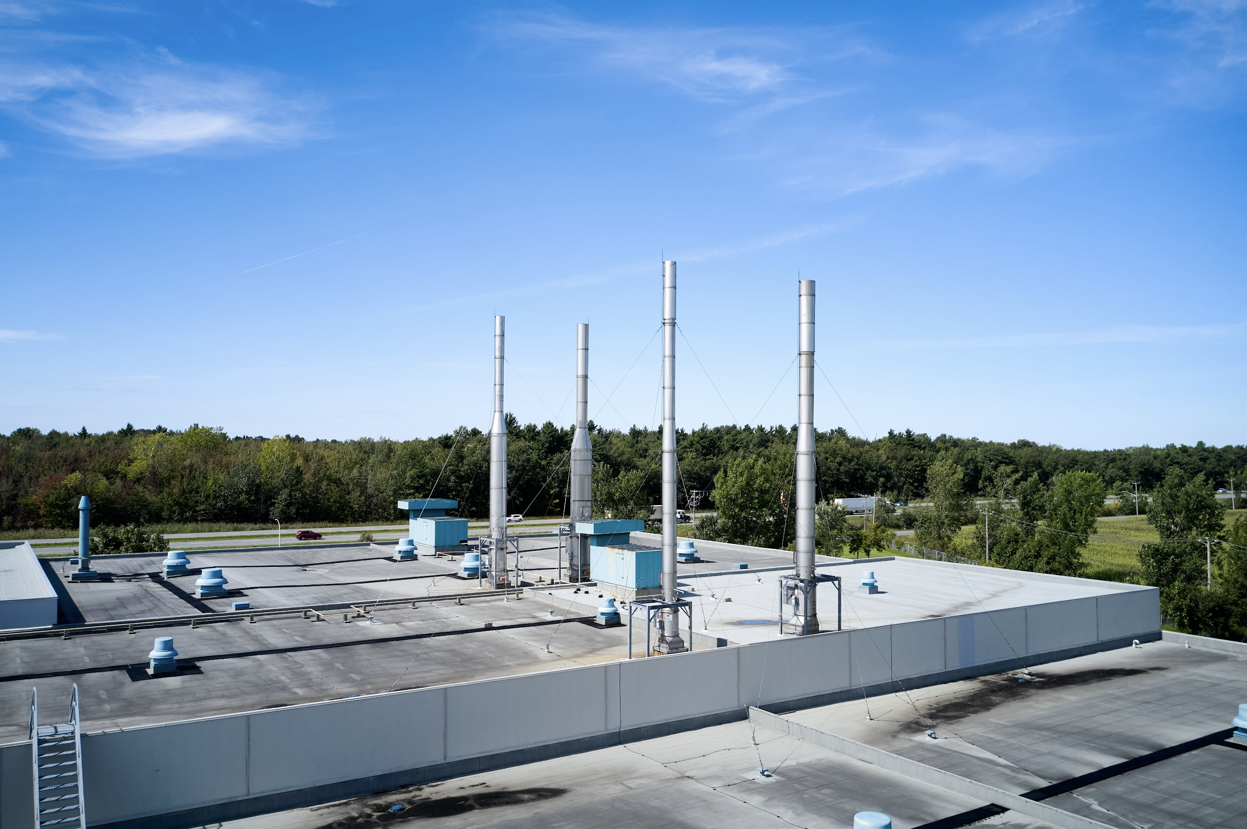 Vue d'une toiture de bâtiment industriel avec plusieurs cheminées et dispositifs de ventilation, entourée d'arbres et un ciel bleu avec quelques nuages.