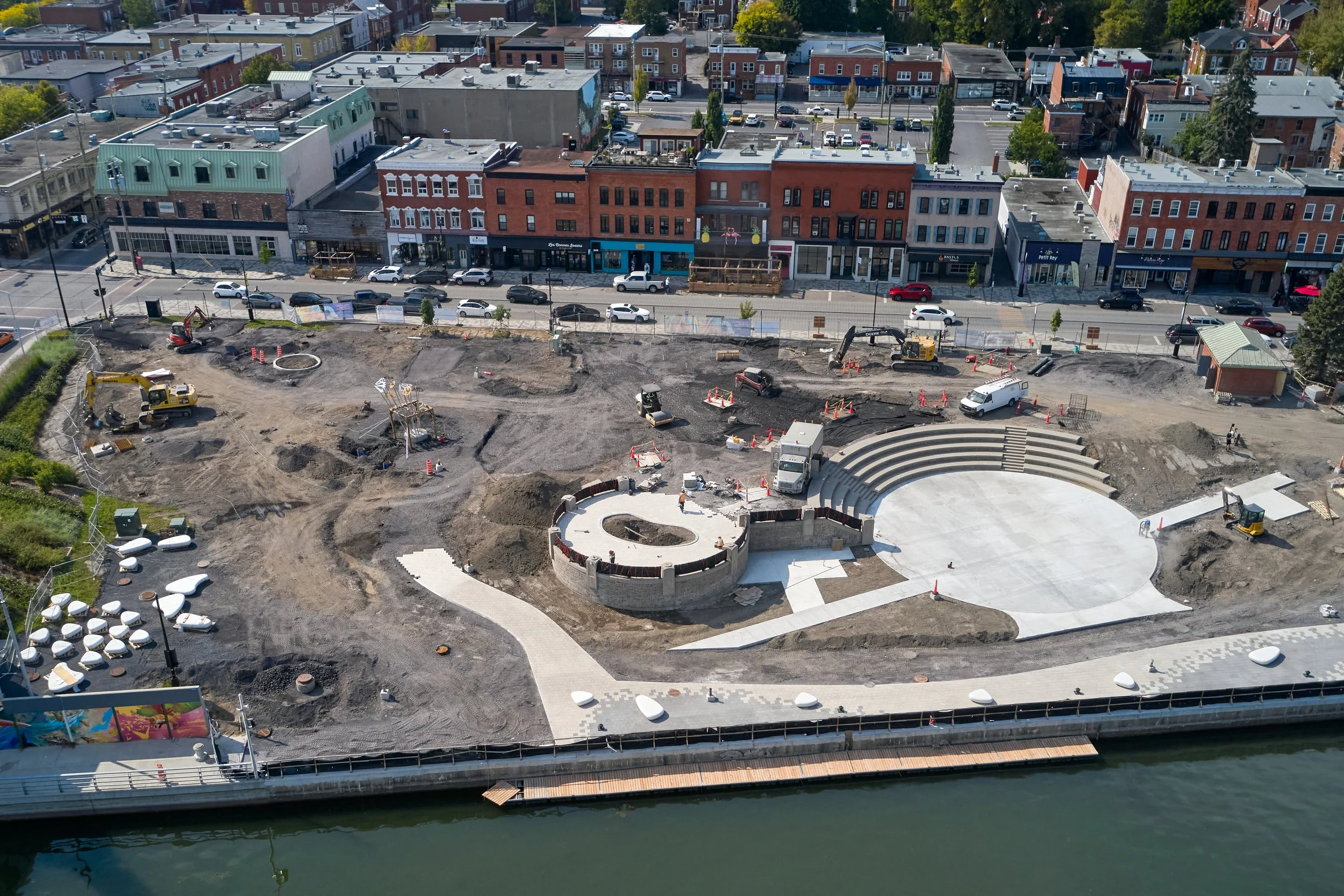 Chantier de construction au bord d'une rivière, avec aménagements paysagers en cours, des engins de chantier, et une zone en béton circulaire et semi-circulaire.