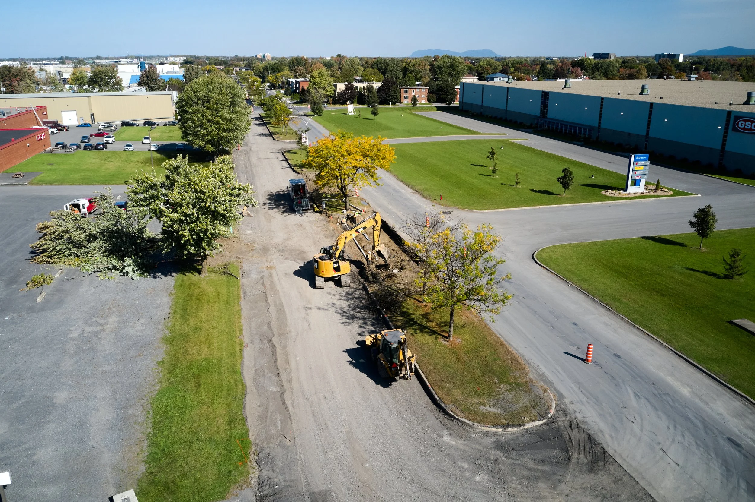 Travaux en cours sur une route avec des machines excavatrices, arbres, espaces verts et un parking à proximité dans une zone industrielle ou commerciale.