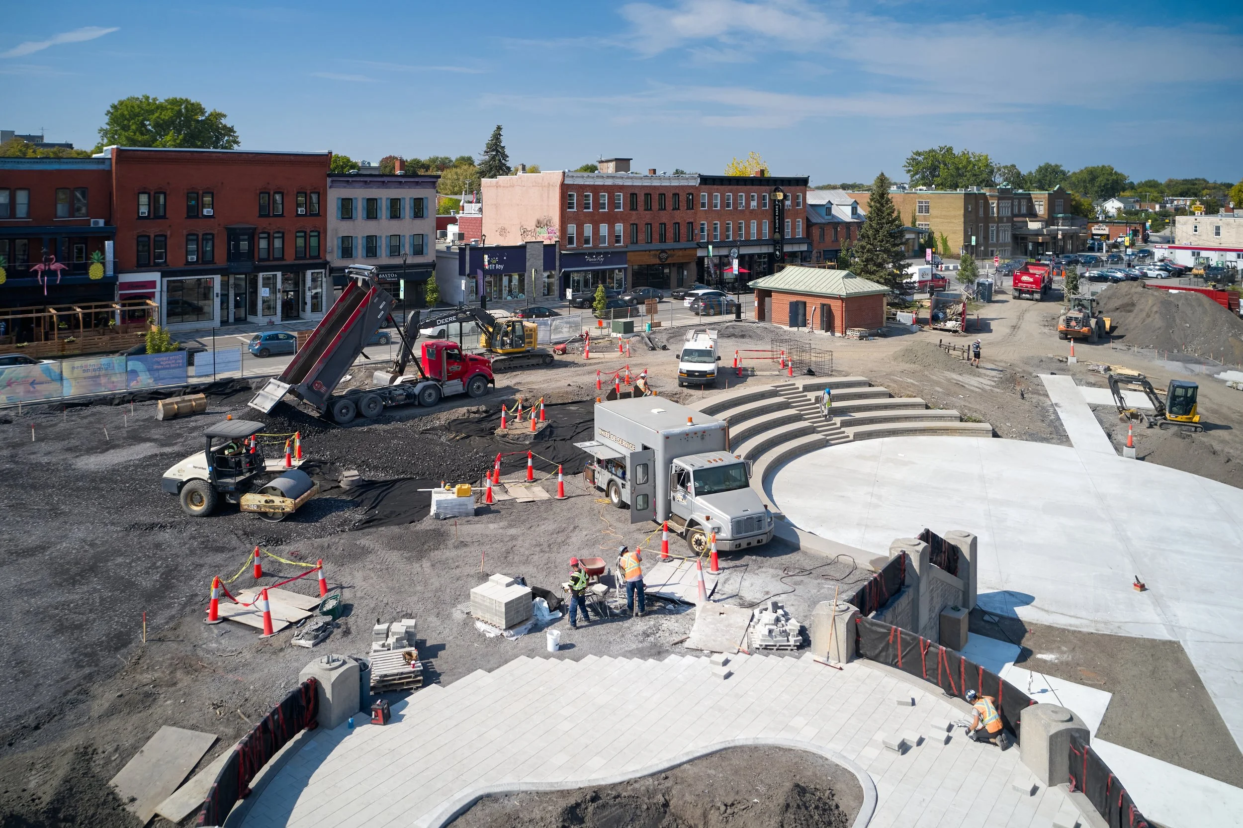 Travaux de construction d'une place publique avec amphithéâtre en béton, plusieurs ouvriers, machines de chantier, et bâtiments commerciaux en arrière-plan.