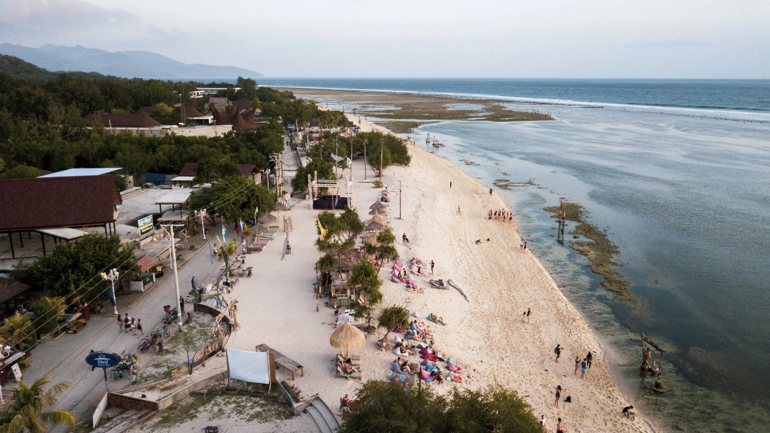 Plage avec des parasols, des personnes allongées au soleil, et des bâtiments le long du rivage, dans un paysage côtier avec de la végétation et des montagnes au loin. Ile des Gilis, Indonésie