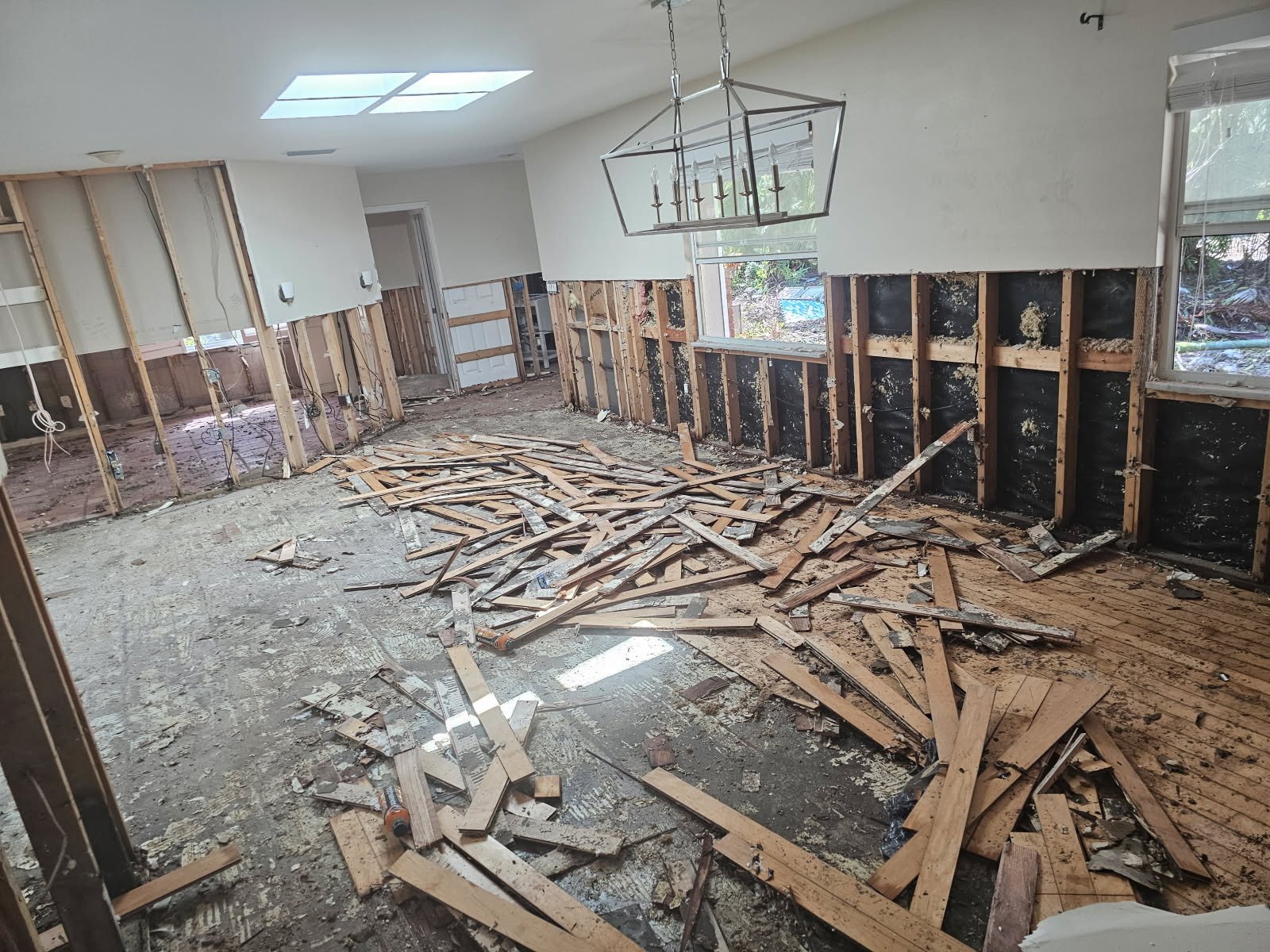 Room under renovation with exposed framing, debris, and removed walls, showcasing construction materials and tools on the floor.