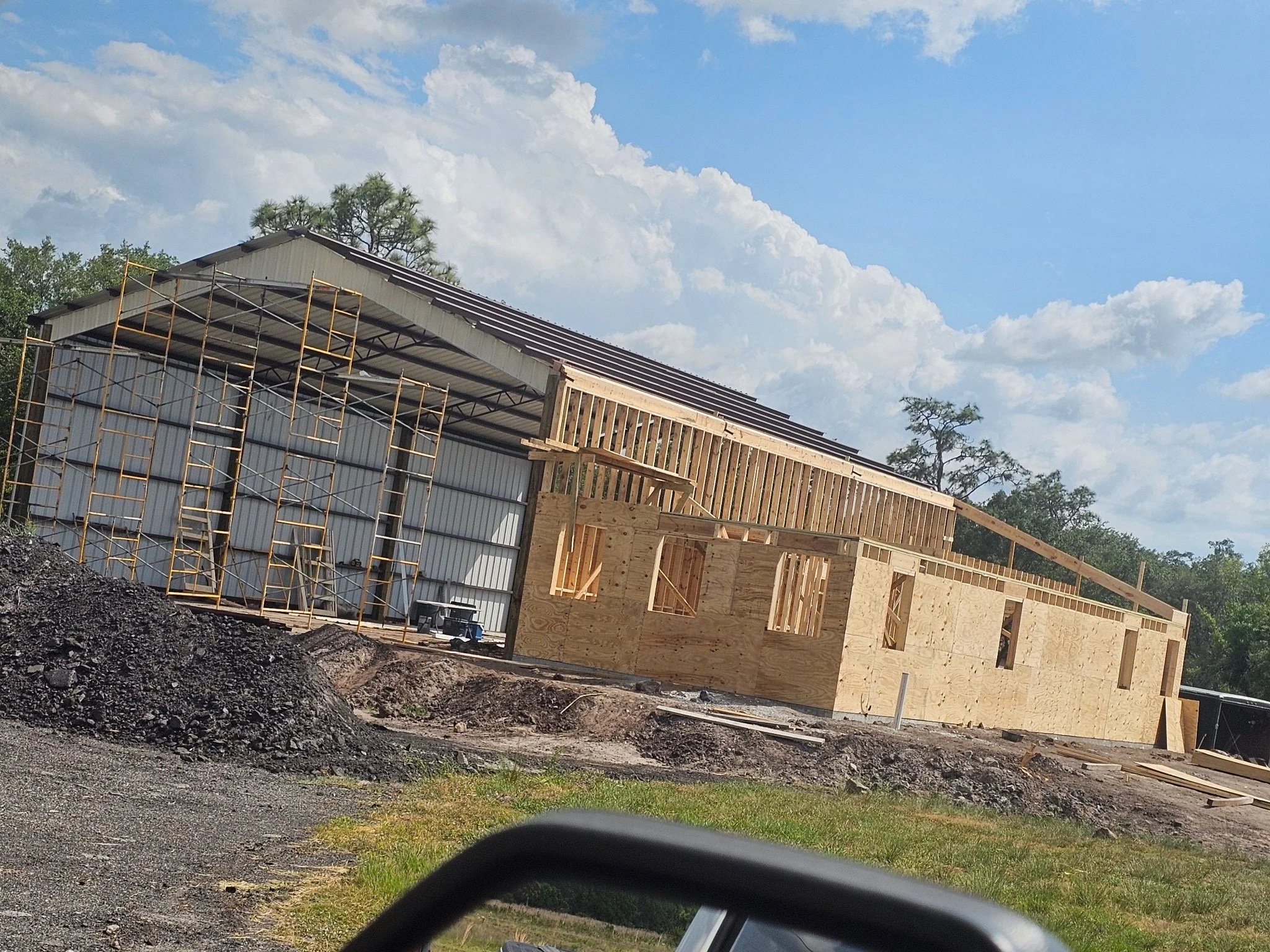 Building under construction with scaffolding and plywood walls, surrounded by dirt and grass, under a blue sky.