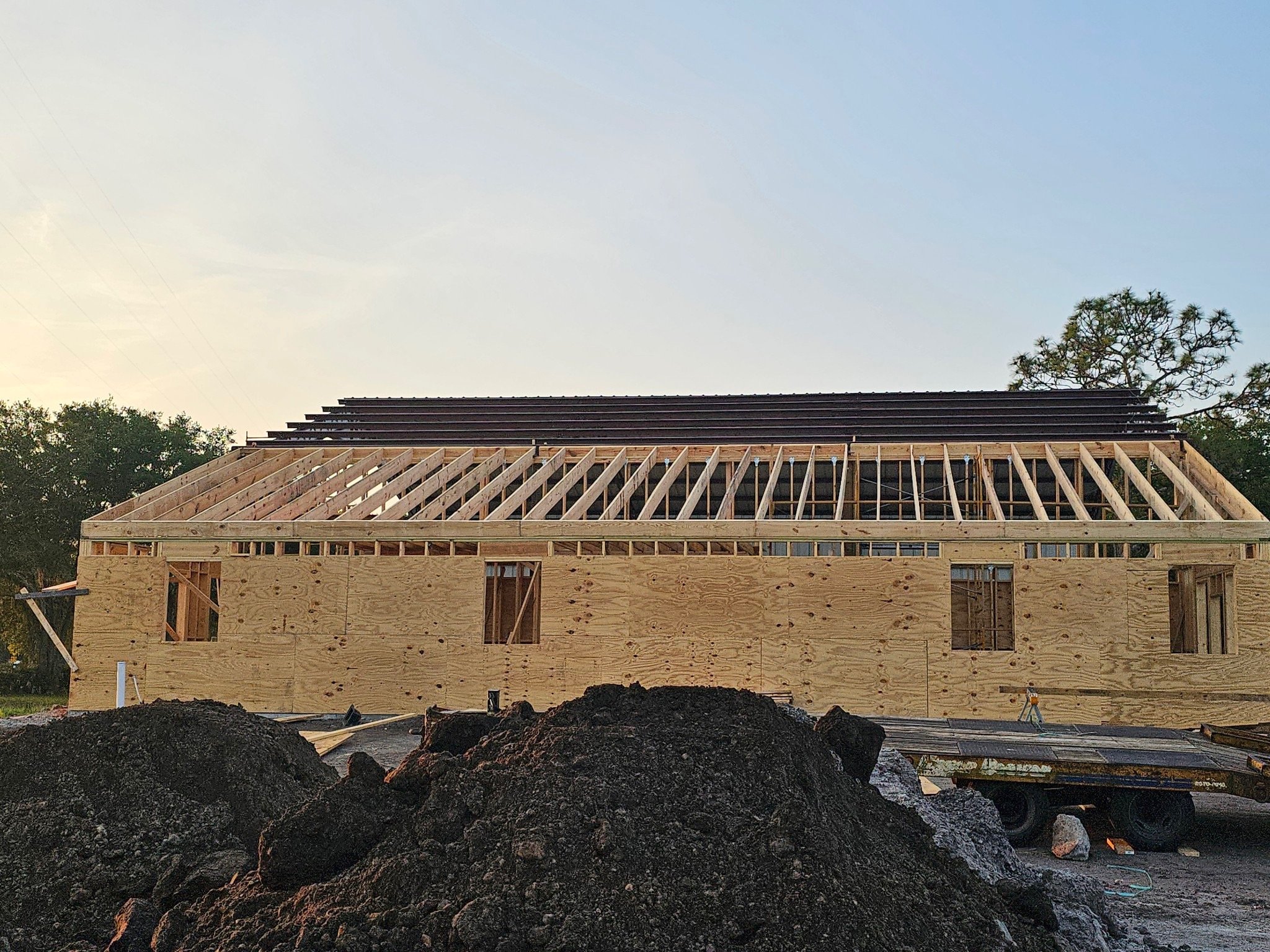Wooden building under construction with roof trusses and plywood walls, surrounded by dirt mounds and trees. PrimeCore builders florida