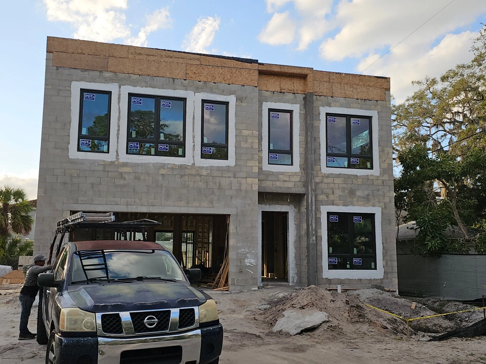 Two-story house under construction with large windows and unfinished exterior, parked truck in front, worker nearby.