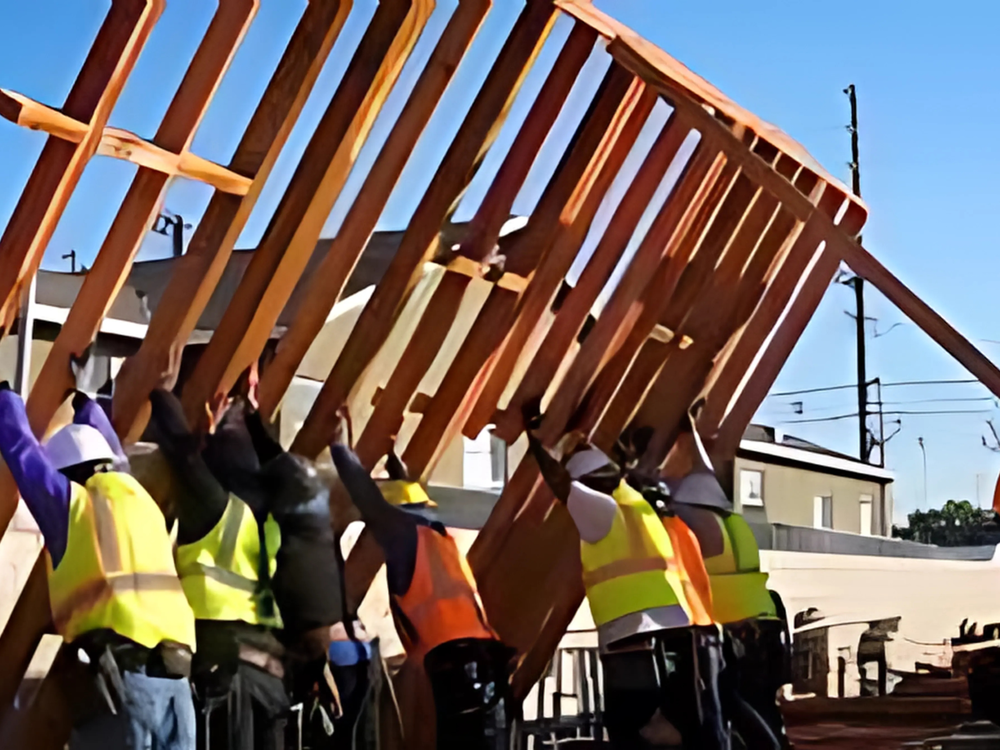Construction workers wearing safety vests and hard hats lifting and positioning a wooden framework against a clear blue sky. PrimeCore builders