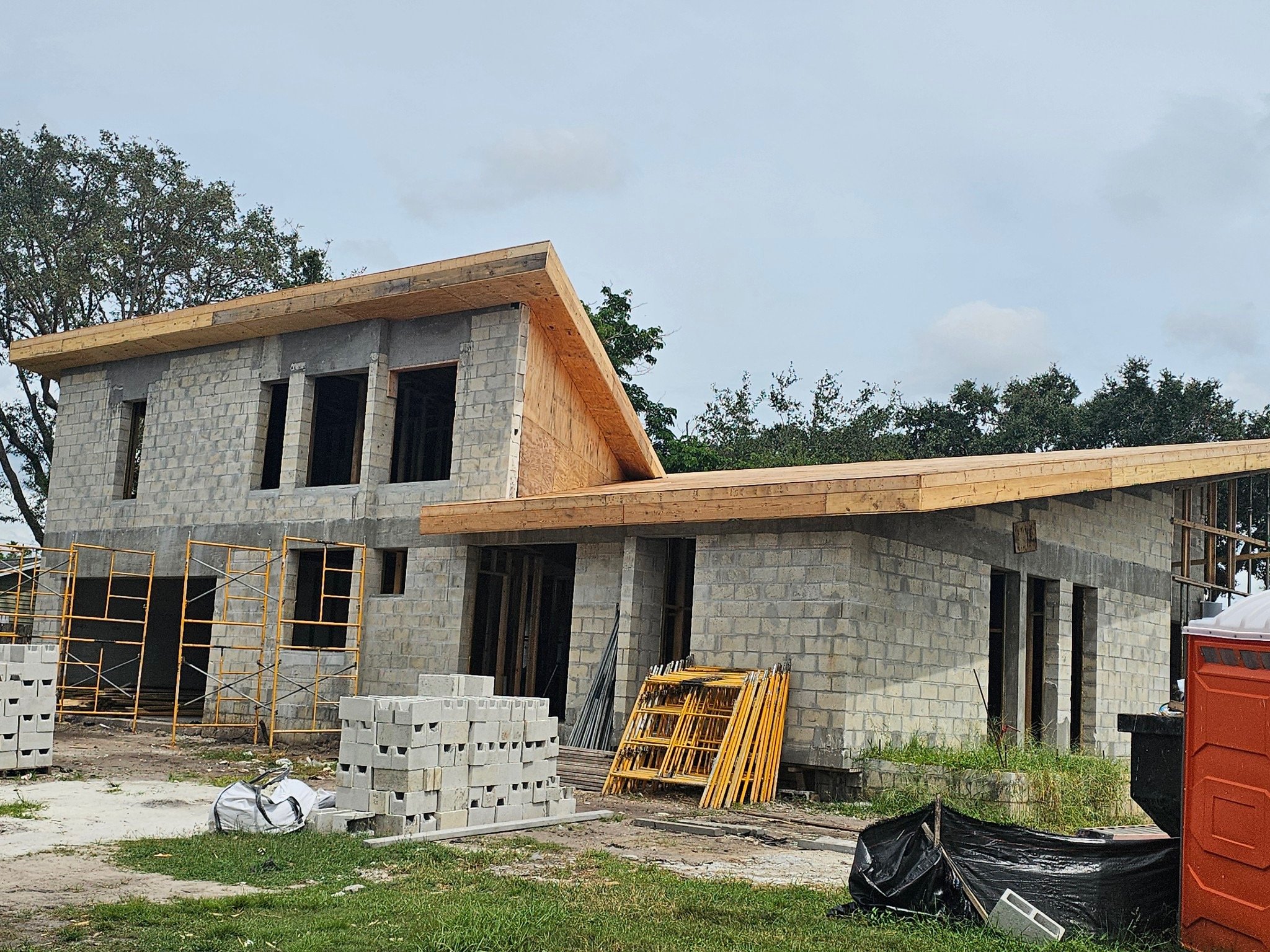A house under construction with a partially completed brick structure, wooden roof framework, scaffolding, and construction materials on site.