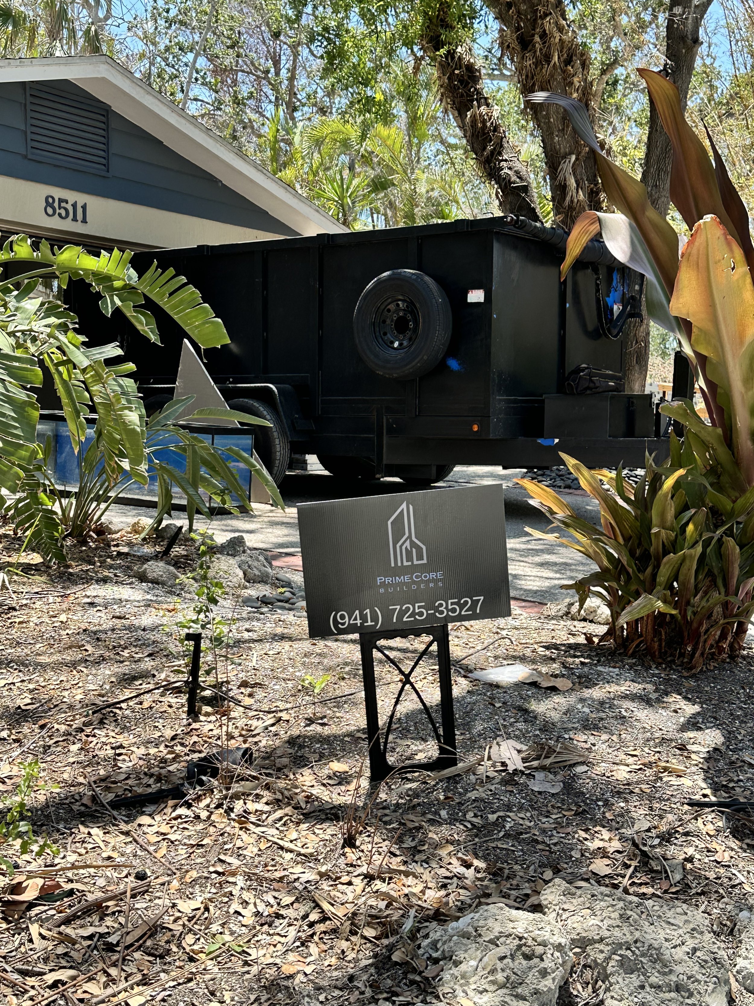 A black trailer parked next to a house surrounded by trees, with a sign in front that reads "Prime Core Builders" and displays a phone number (941) 725-3527. The house number 8511 is visible above the trailer. The ground is covered in dry leaves and