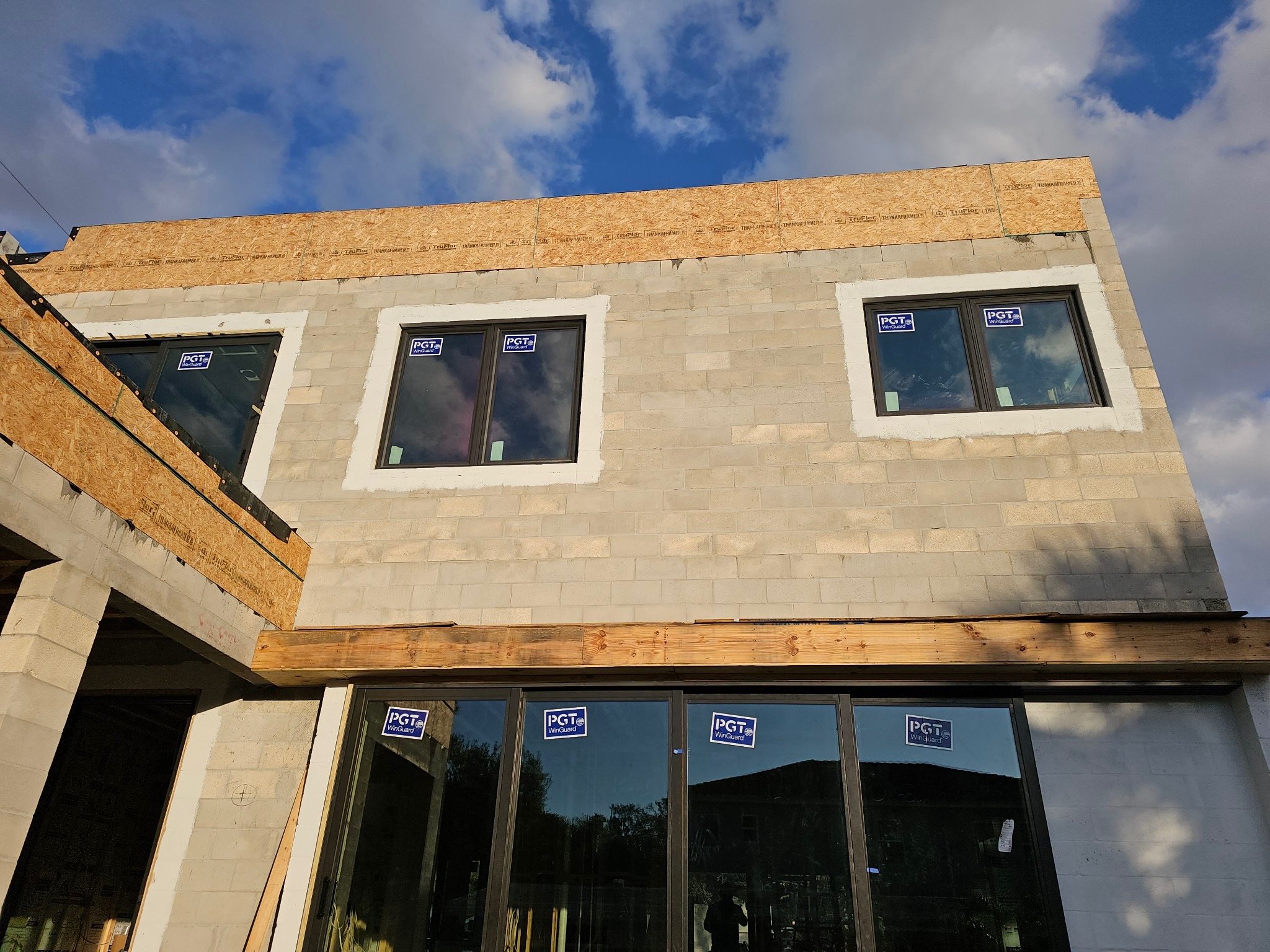 A partially constructed building with gray concrete blocks and wood framing. It features multiple PGT-branded windows with stickers, and a cloudy sky in the background. PrimeCore builders