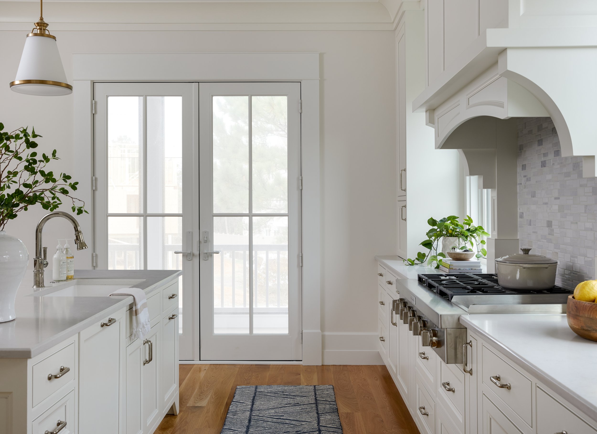 Modern kitchen with white cabinets, stainless steel appliances, a large gas stove, potted plants, and a double French door leading outside. The room is bright with natural light and features a wooden floor and white decor.