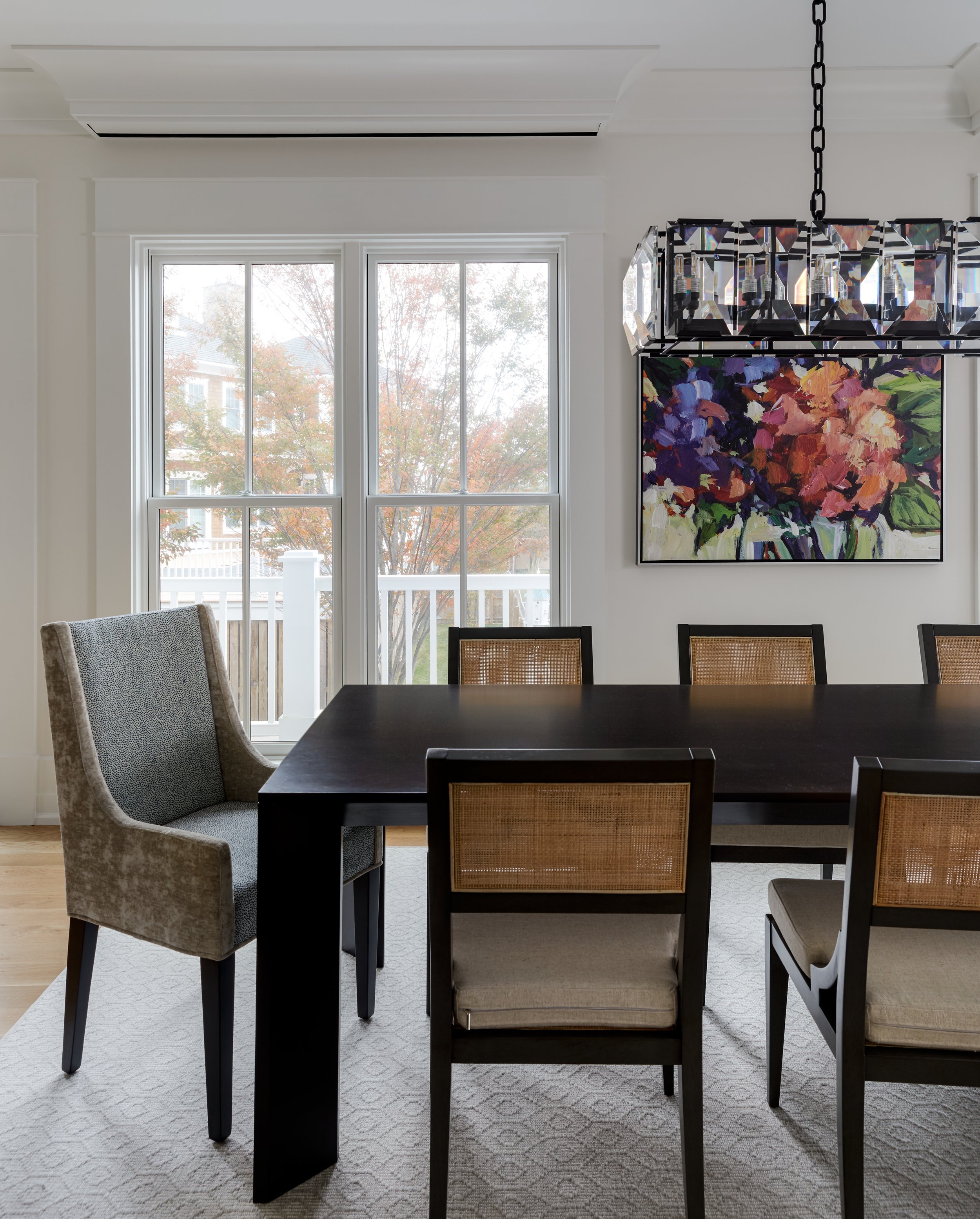 Modern dining room with a dark wooden table and six chairs, large window, colorful abstract painting, and a crystal chandelier.