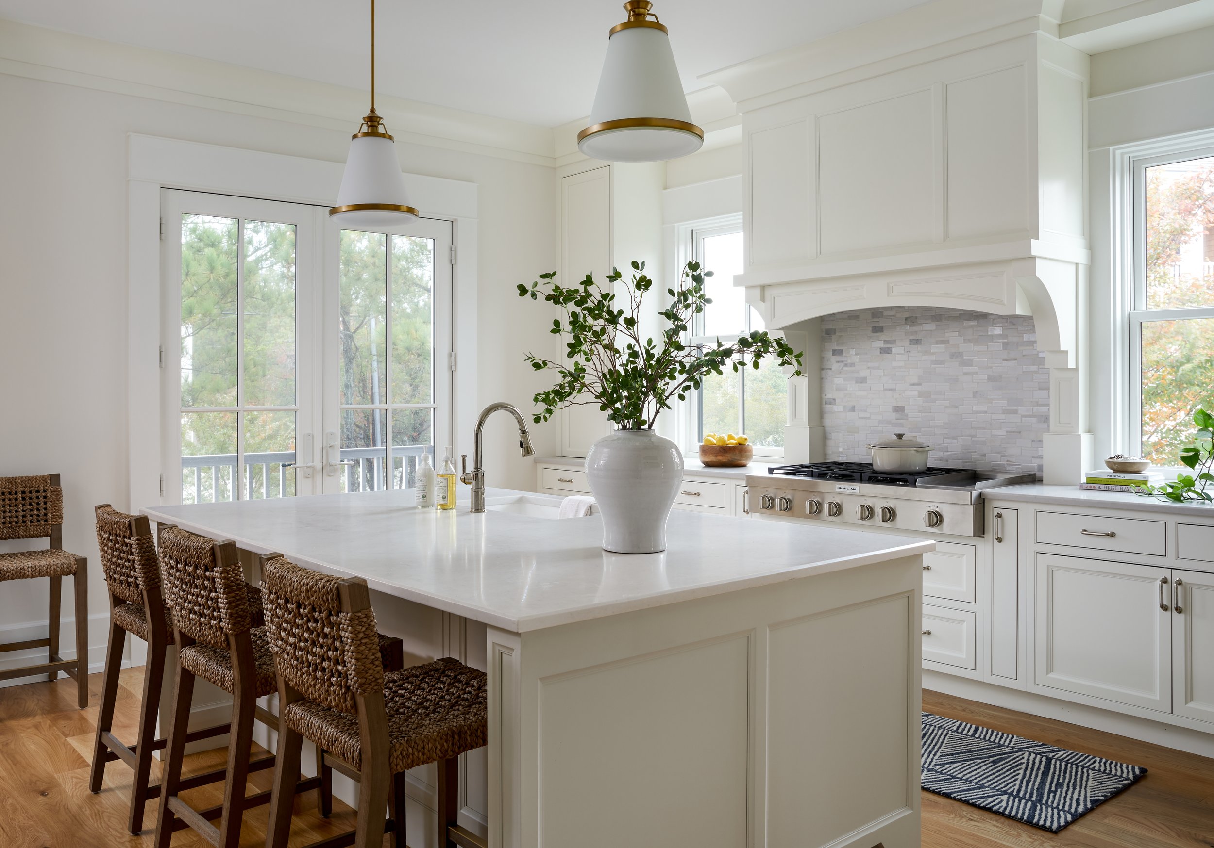 Modern kitchen with white cabinets, marble countertop, woven barstools, pendant lighting, vase with greenery, stove with a pot, and large windows with a garden view.