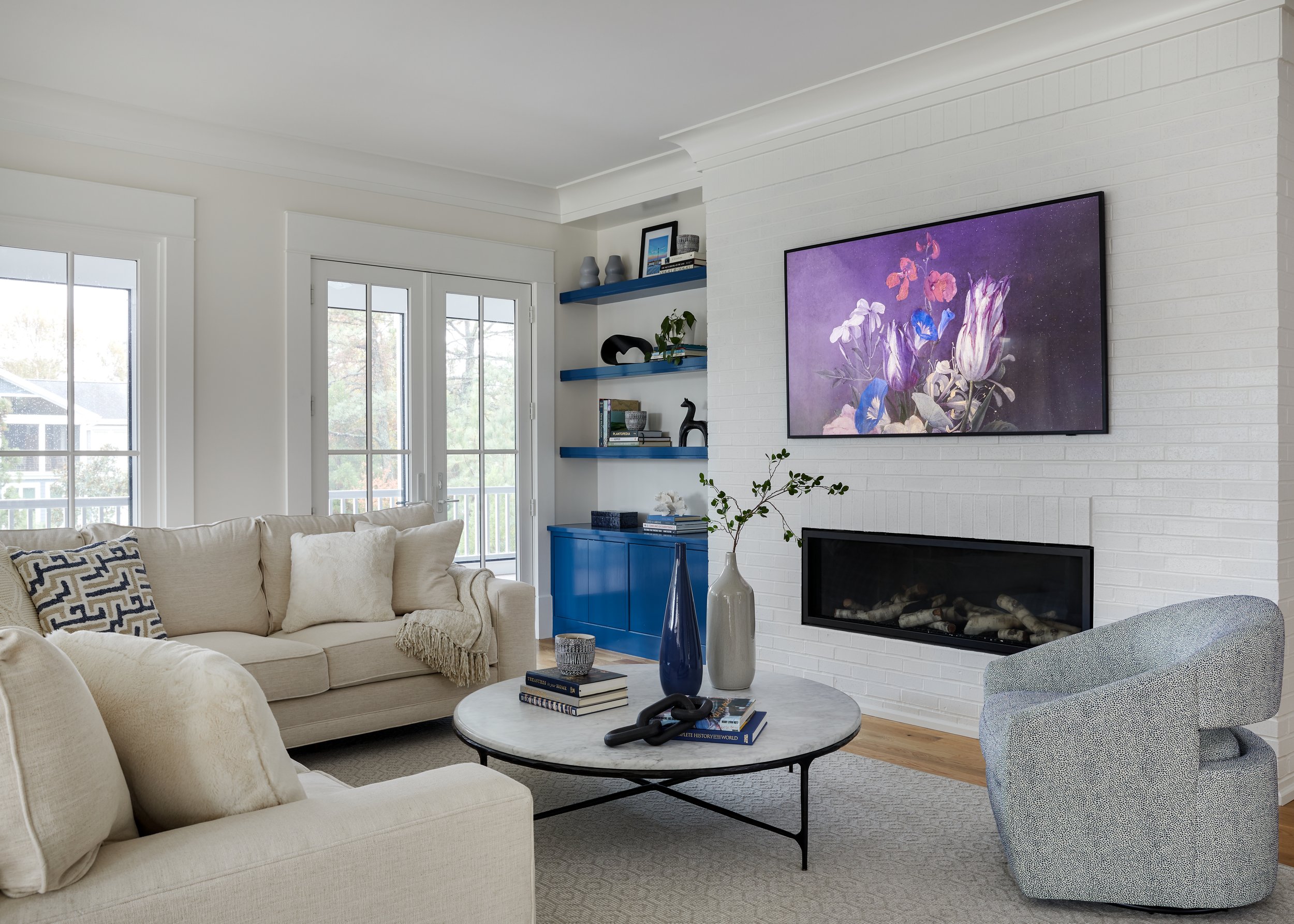 Modern living room with light beige sofas, round marble coffee table with decor, blue accent shelves, wall art, and large windows.