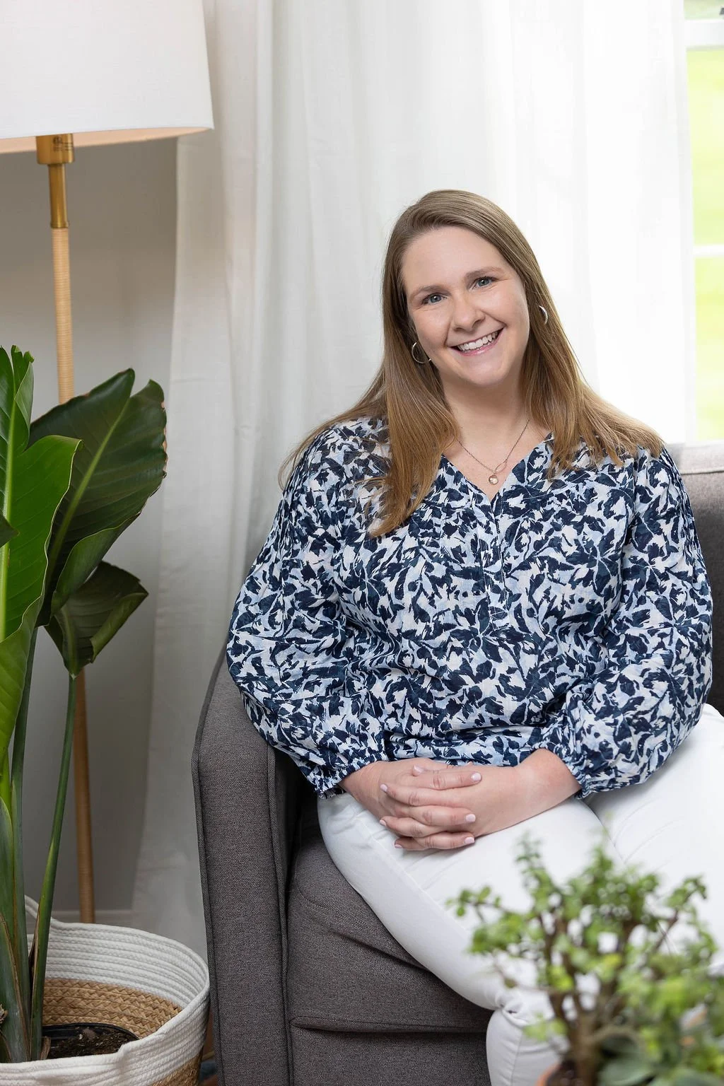 A smiling and empathic woman (Dr. Cathy Bykowski) with shoulder-length light brown hair, wearing a blue and white patterned blouse, sitting on a gray chair in a bright room with white curtains and green plants.