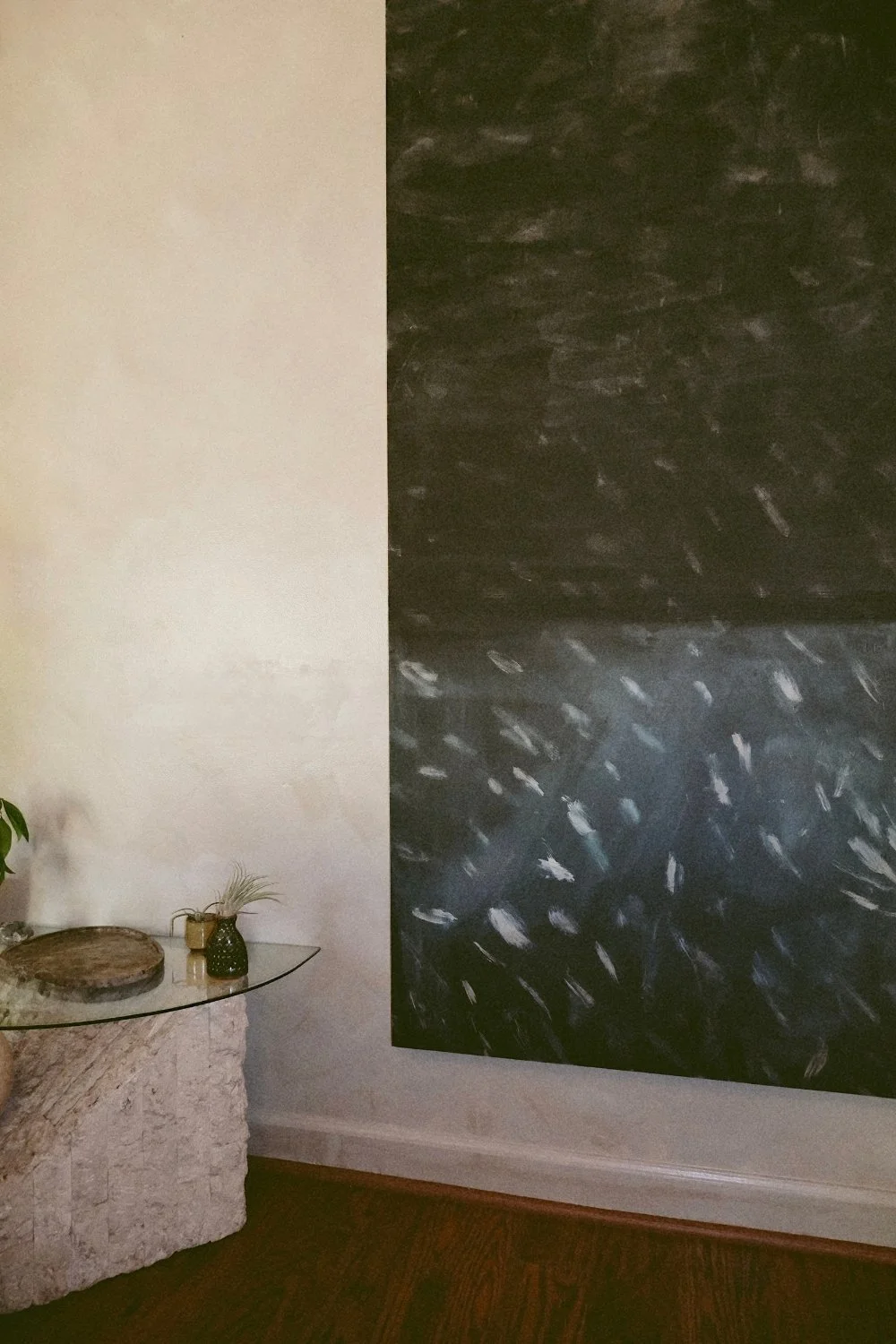 A room corner with a textured black and dark gray abstract wall art, a small round stone table with a glass top, a dark green vase with a plant, and a small light-colored vase on the table.