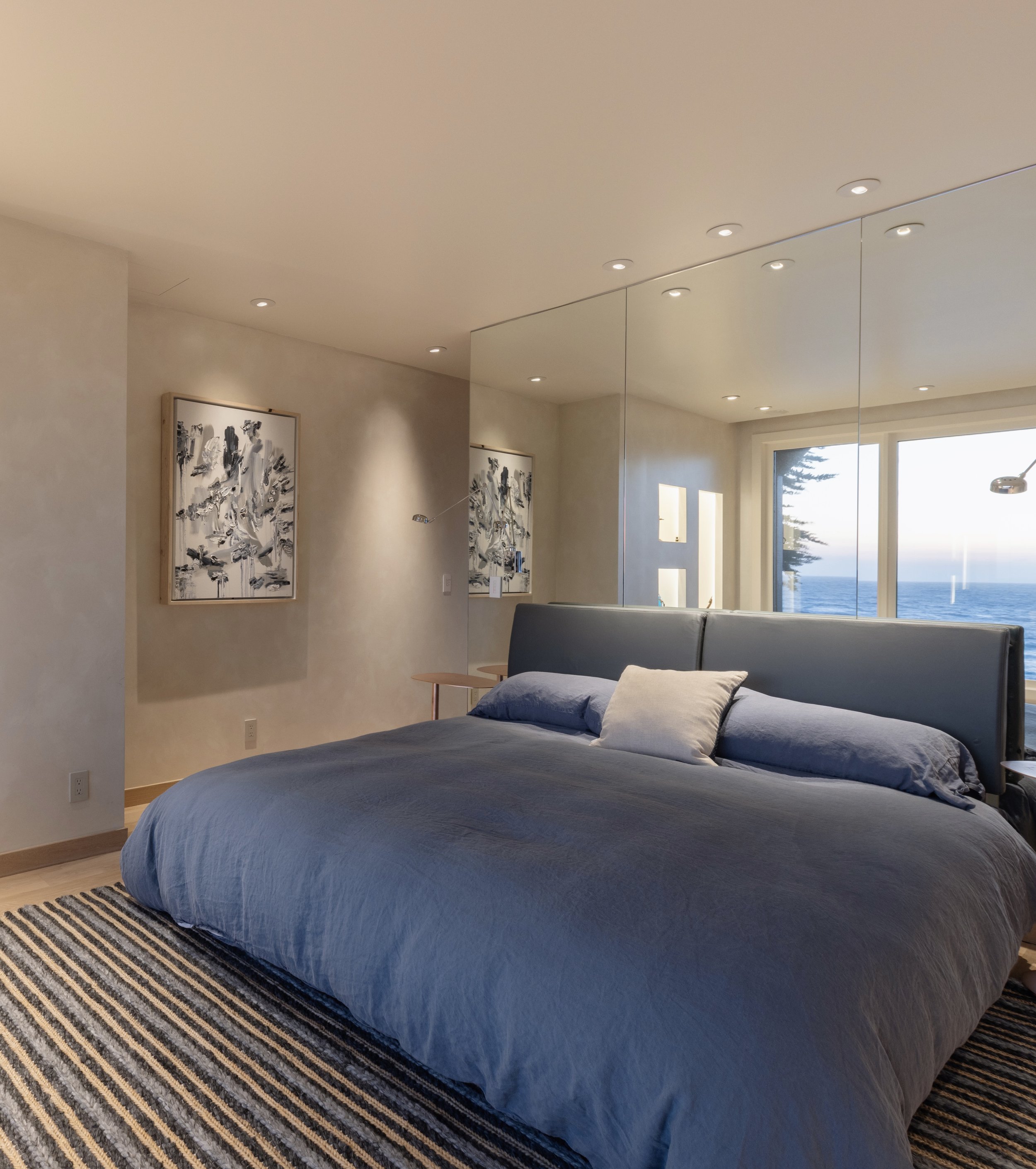 Modern bedroom with a large bed featuring a blue comforter, a white pillow, and a striped rug. A large mirror wall reflects the ocean view visible through a window. Two abstract paintings are hung on the walls.