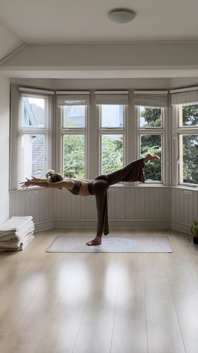 Person practicing yoga in a room with large windows, performing a standing balance pose on a yoga mat.