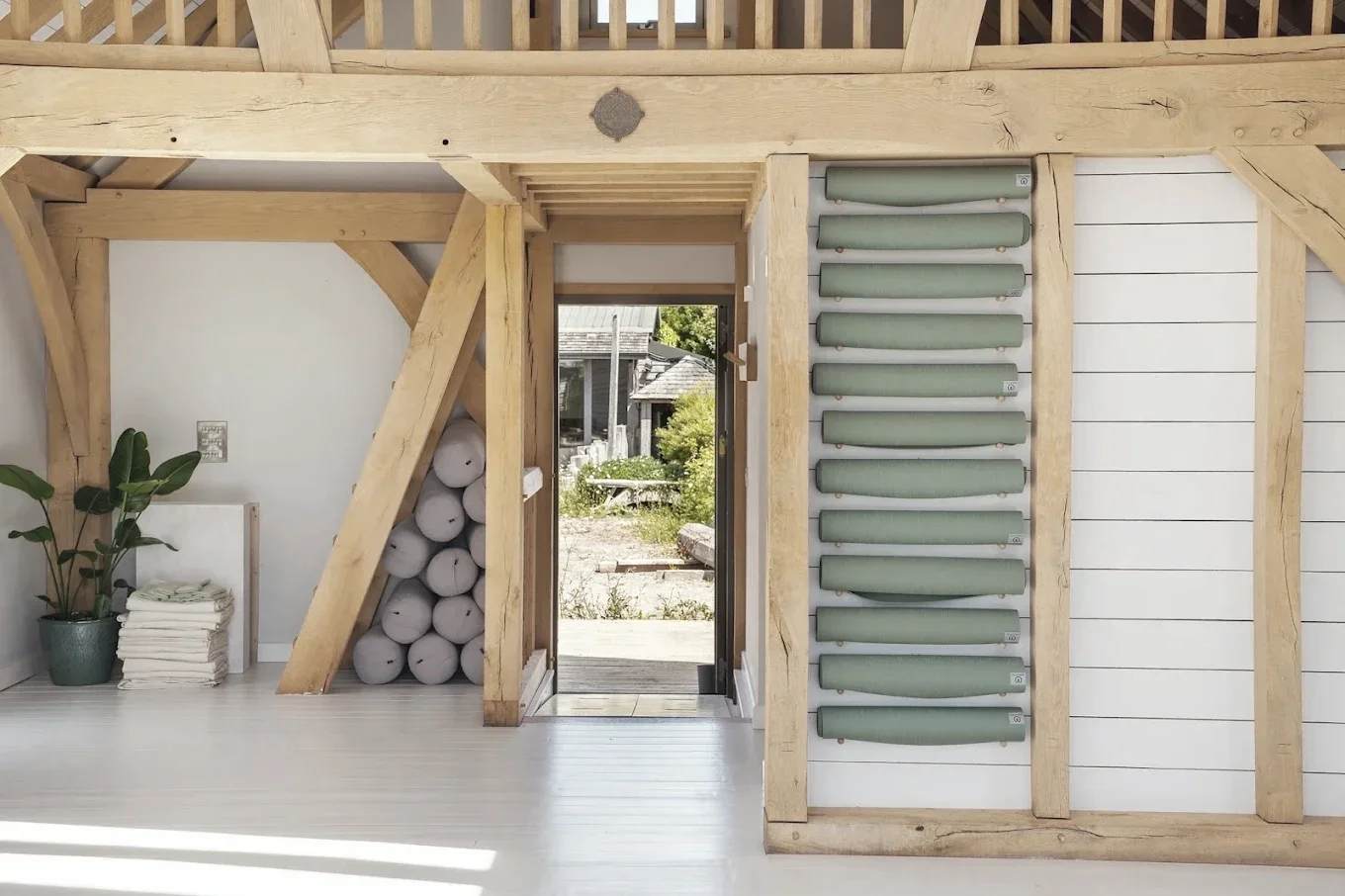 Interior of a minimally decorated room with exposed wooden beams, a potted plant, neatly stacked folded towels, and a wall with rolled-up yoga mats, leading to an outdoor area with trees and a small building.